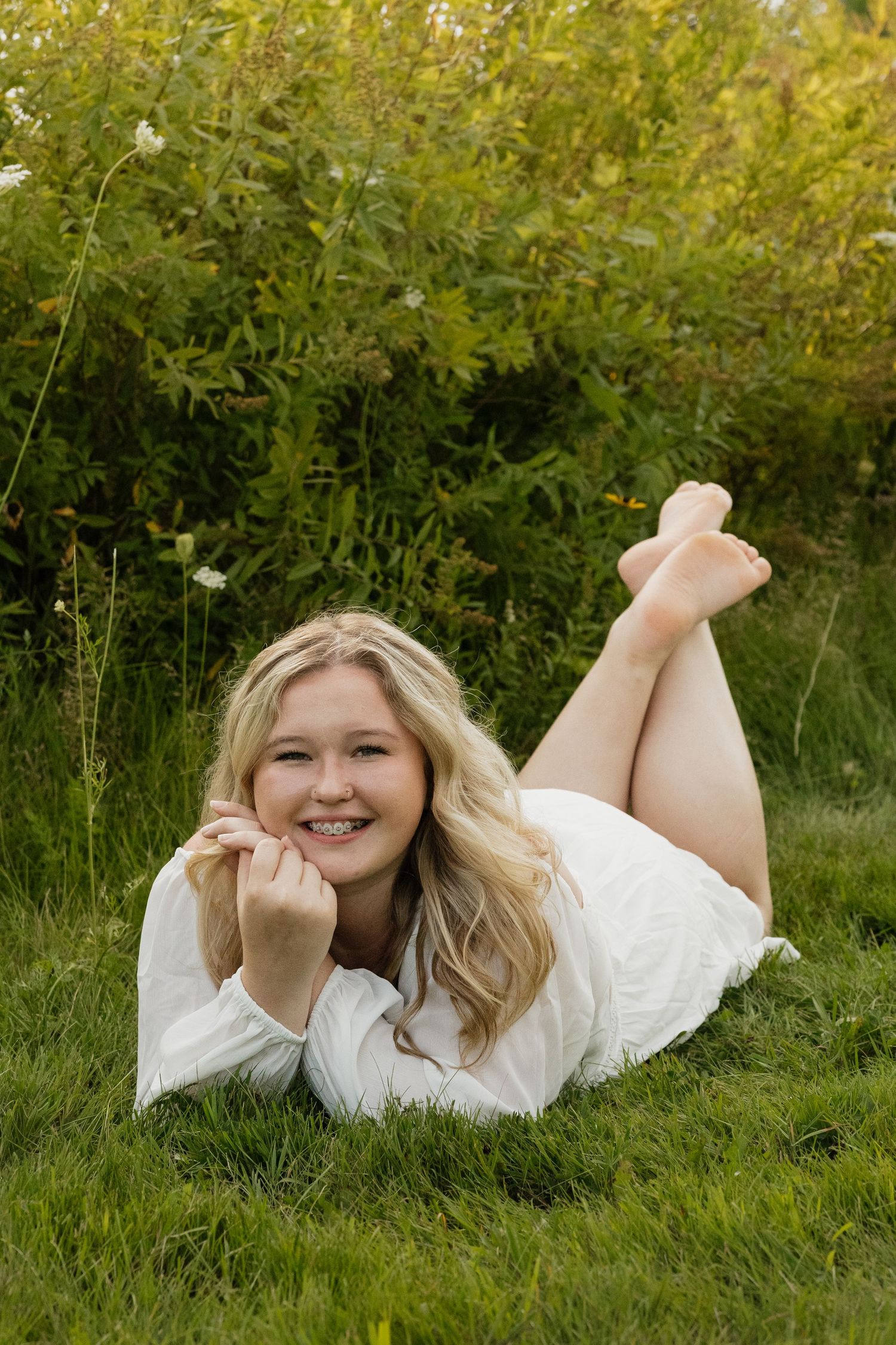 Smiling blonde teen in white dress lying on green grass with legs crossed behind her outdoors.
