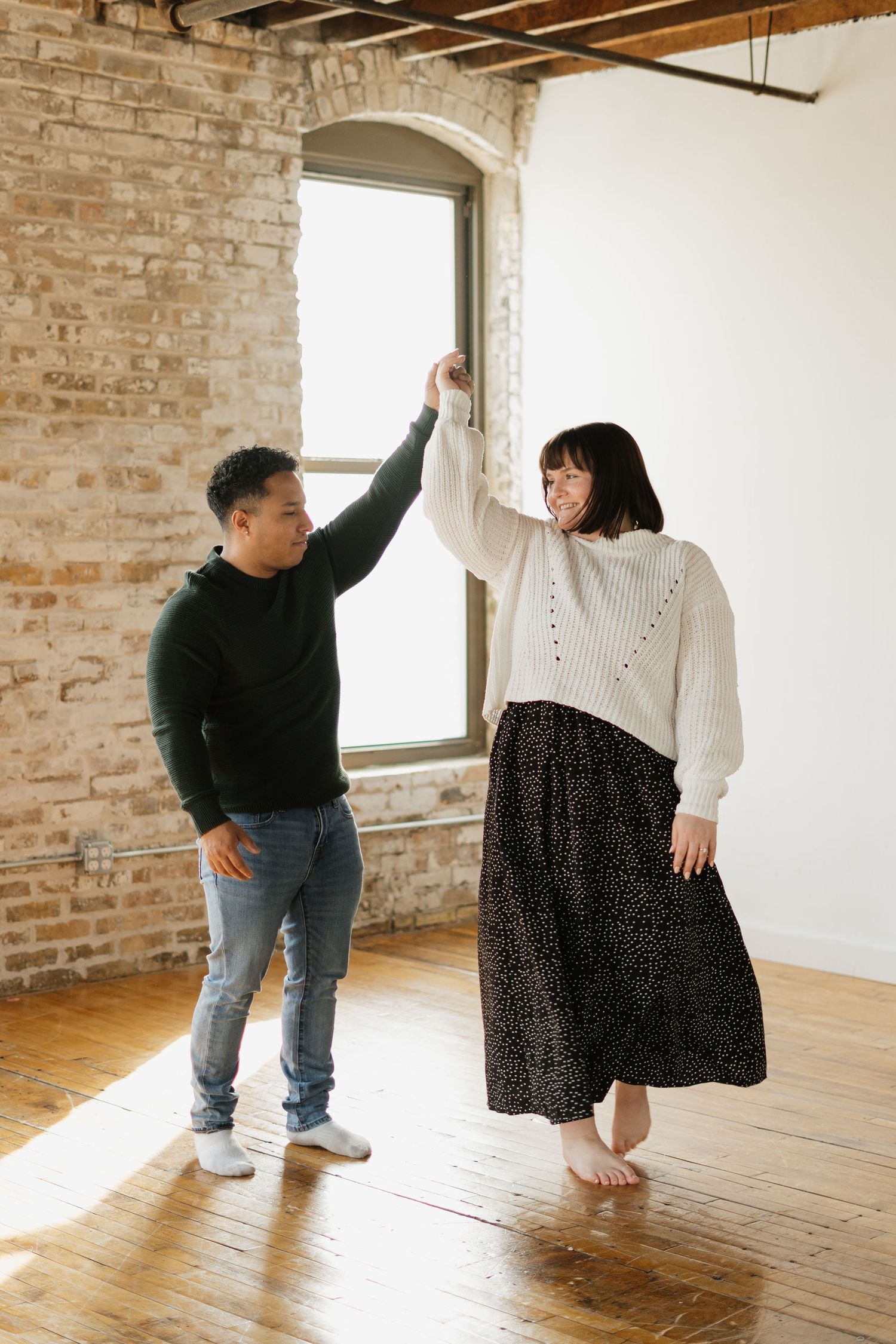 A couple dances joyfully in a bright loft space with exposed brick walls and hardwood floors.