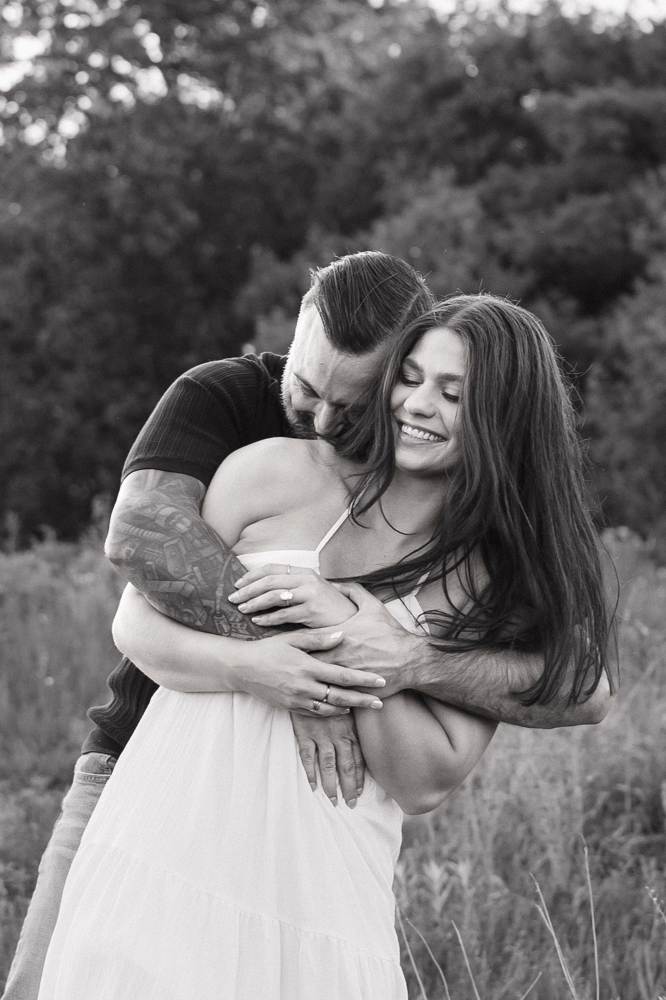 Black and white photo of a tattooed man embracing a smiling woman in a white dress outdoors.