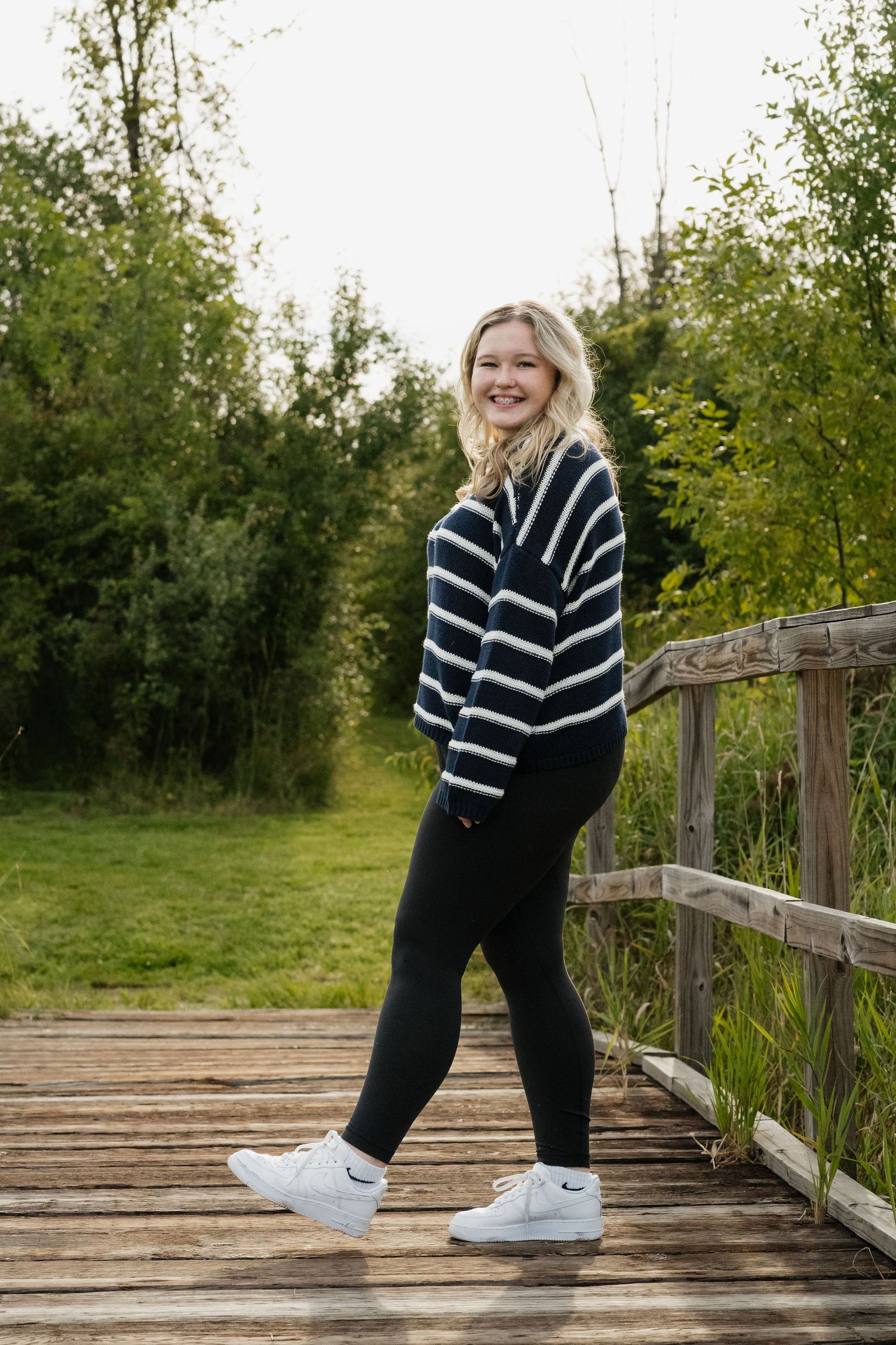 Smiling blonde teen in striped sweater and black leggings standing on a wooden bridge surrounded by greenery.