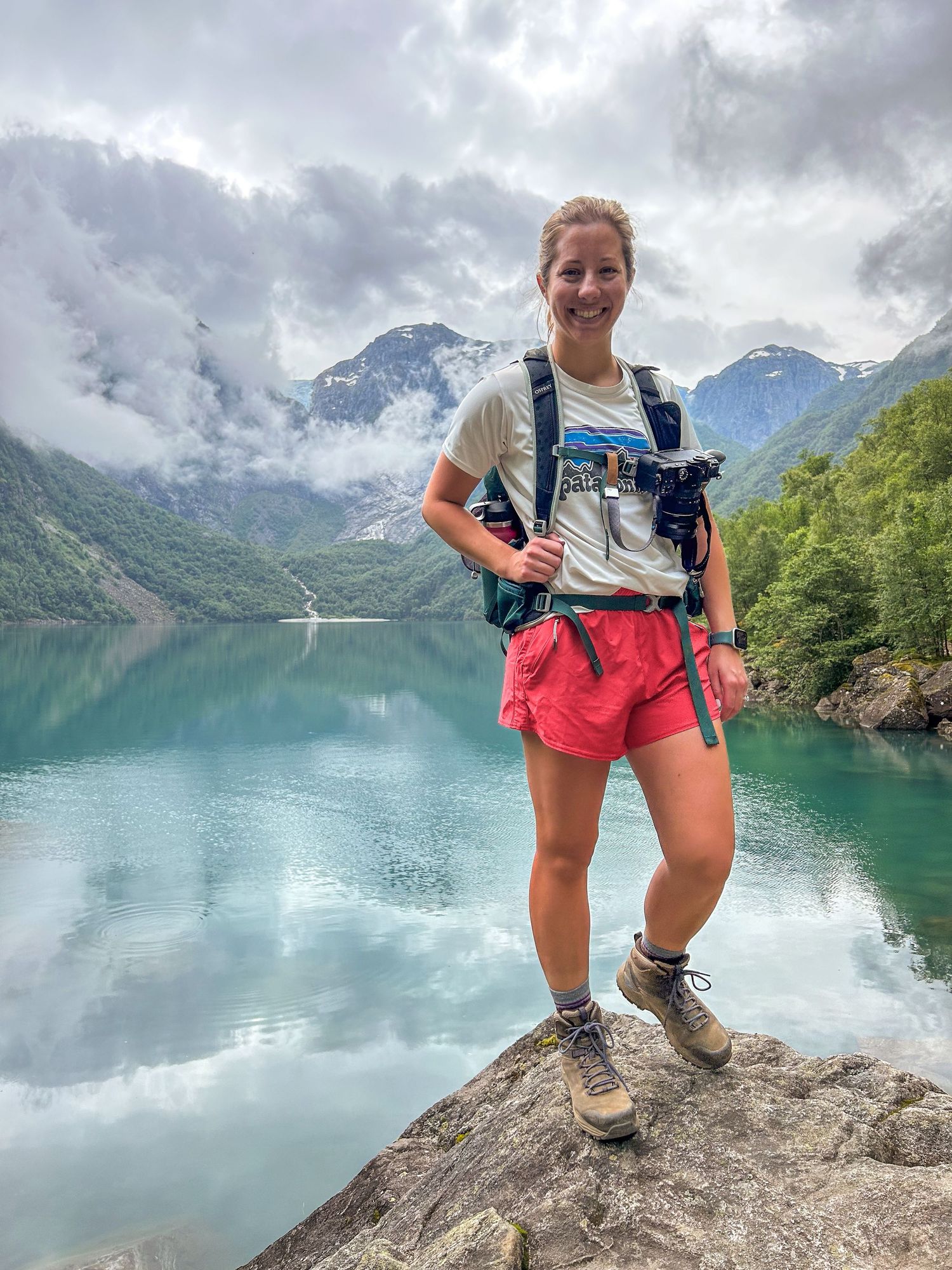 Smiling female hiker standing on rock with stunning turquoise mountain lake and cloudy peaks behind her.