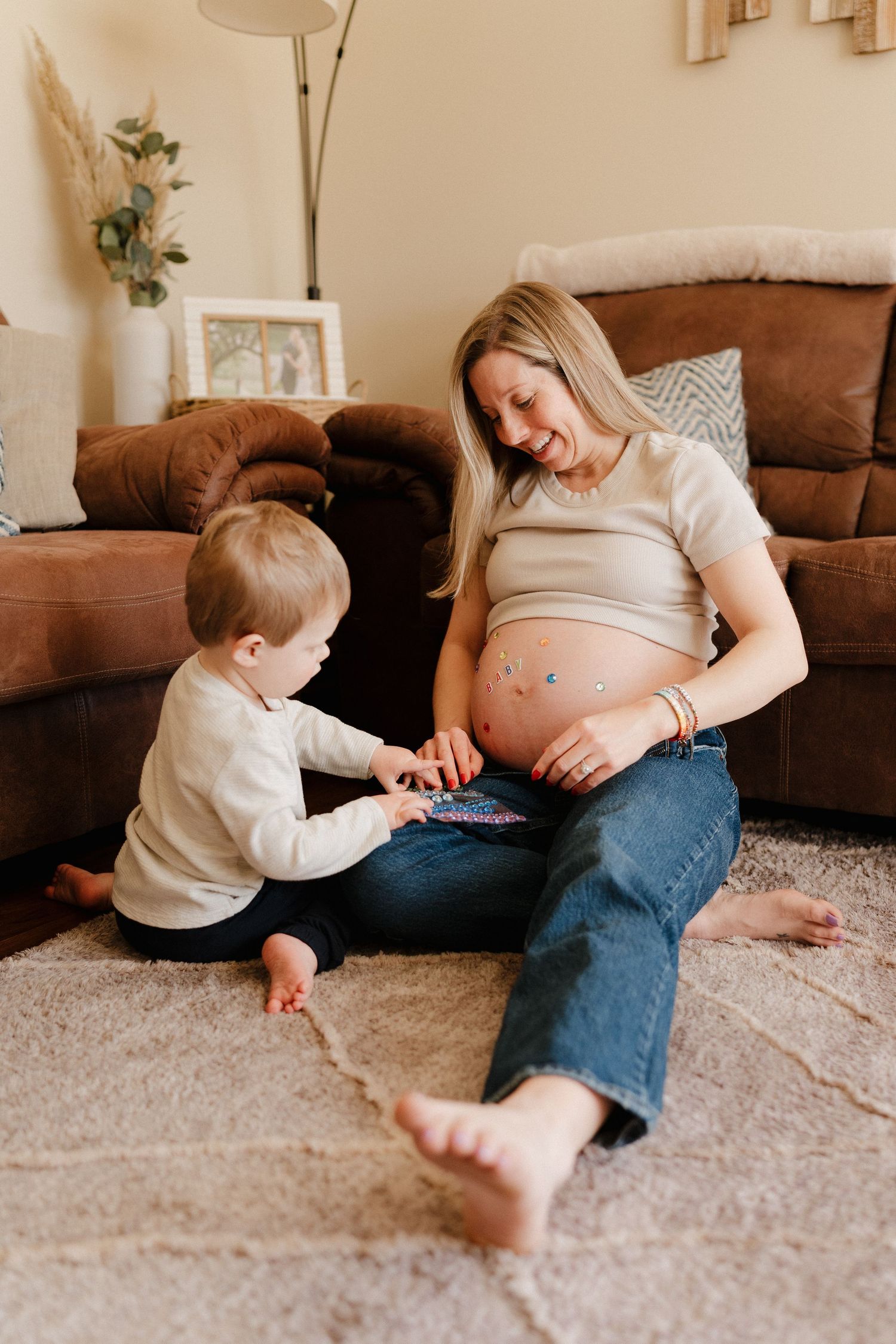Pregnant woman sitting on floor with toddler placing stickers on her baby bump in cozy living room.