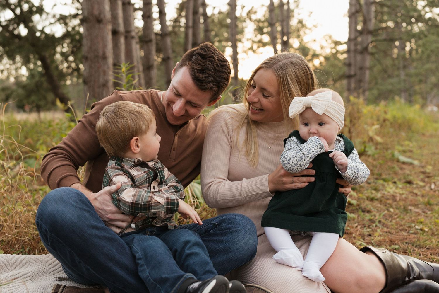 Family of four sits on blanket in pine forest, parents smiling at baby girl held by mother.