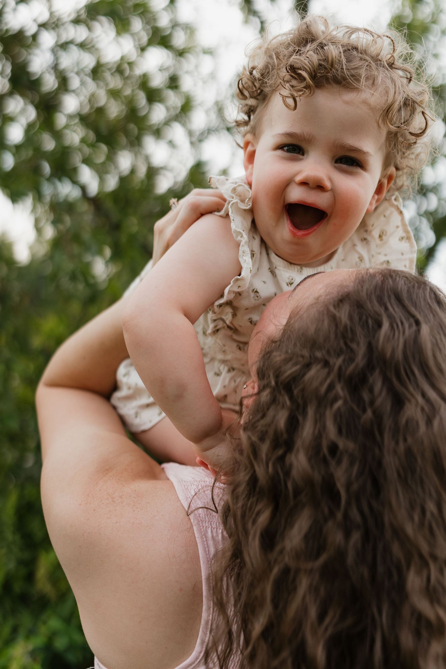 A laughing toddler with curly hair is lifted up by her mother in a lush green outdoor setting.