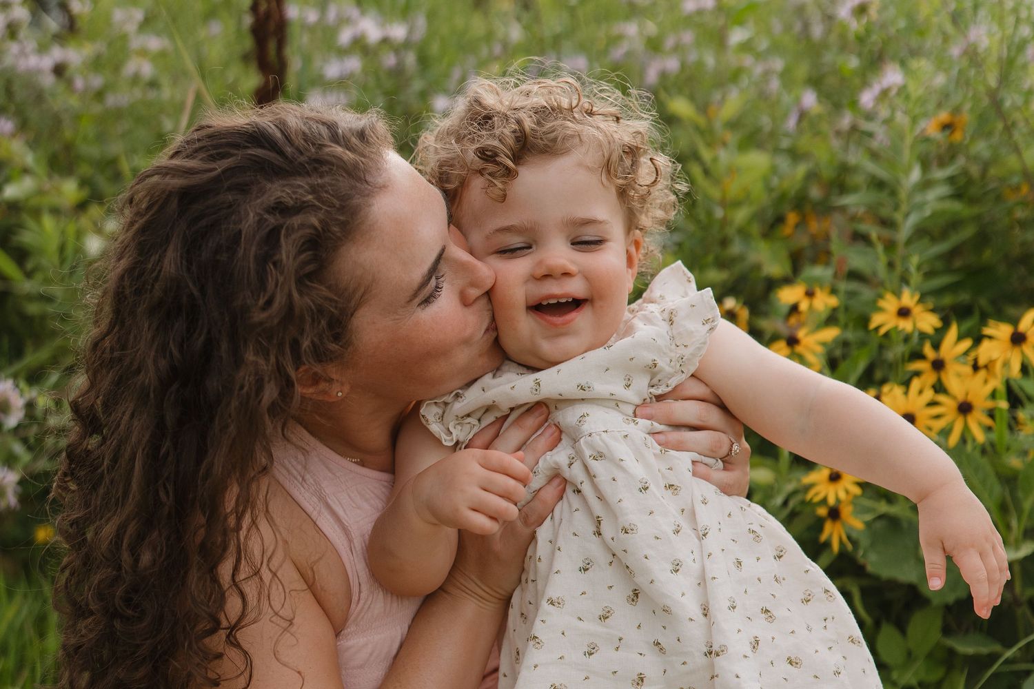 A mother kisses her laughing toddler girl with curly hair in a field of wildflowers.