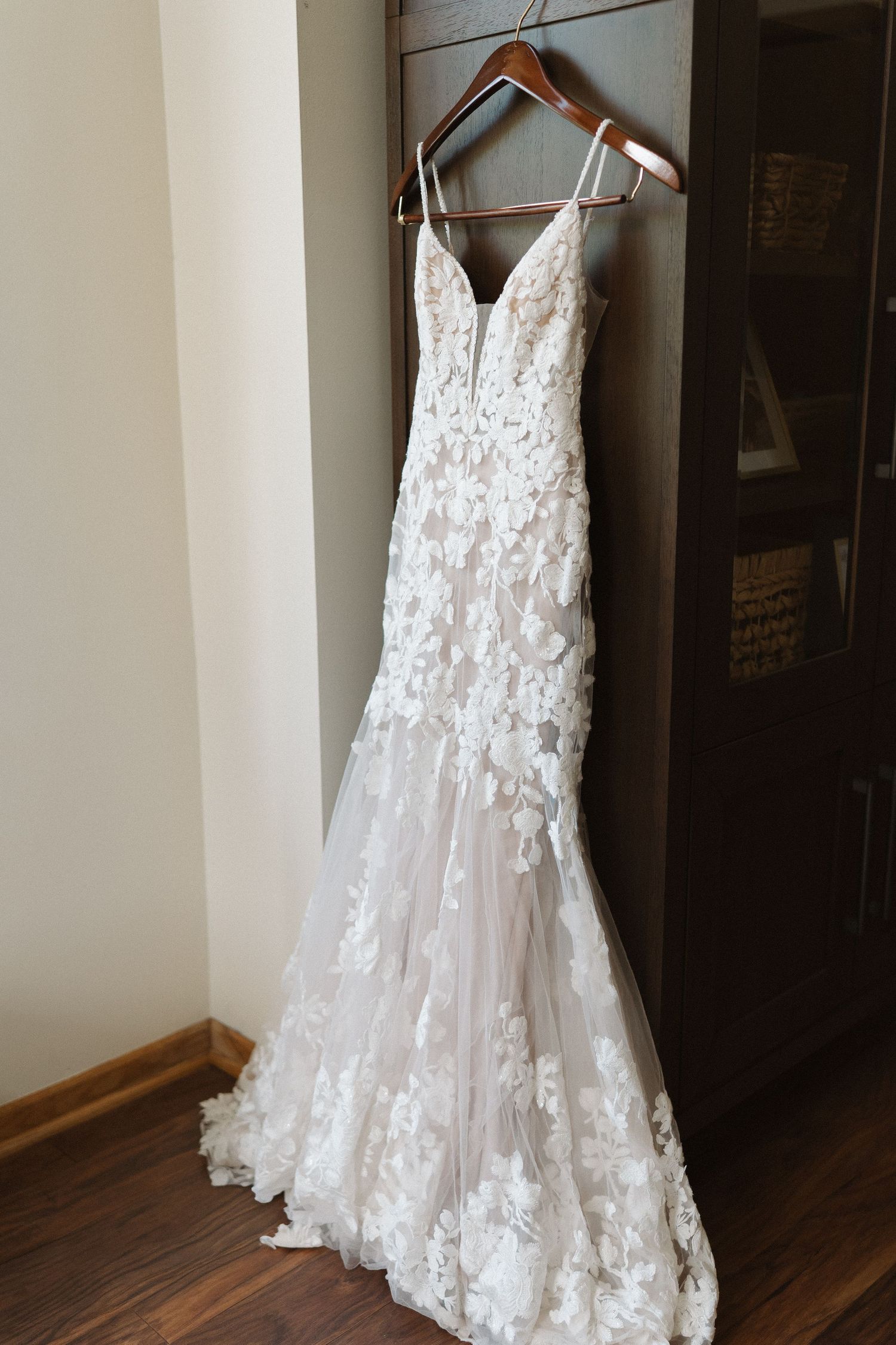 Elegant white lace mermaid wedding dress with spaghetti straps hanging on a wooden hanger against a neutral wall.