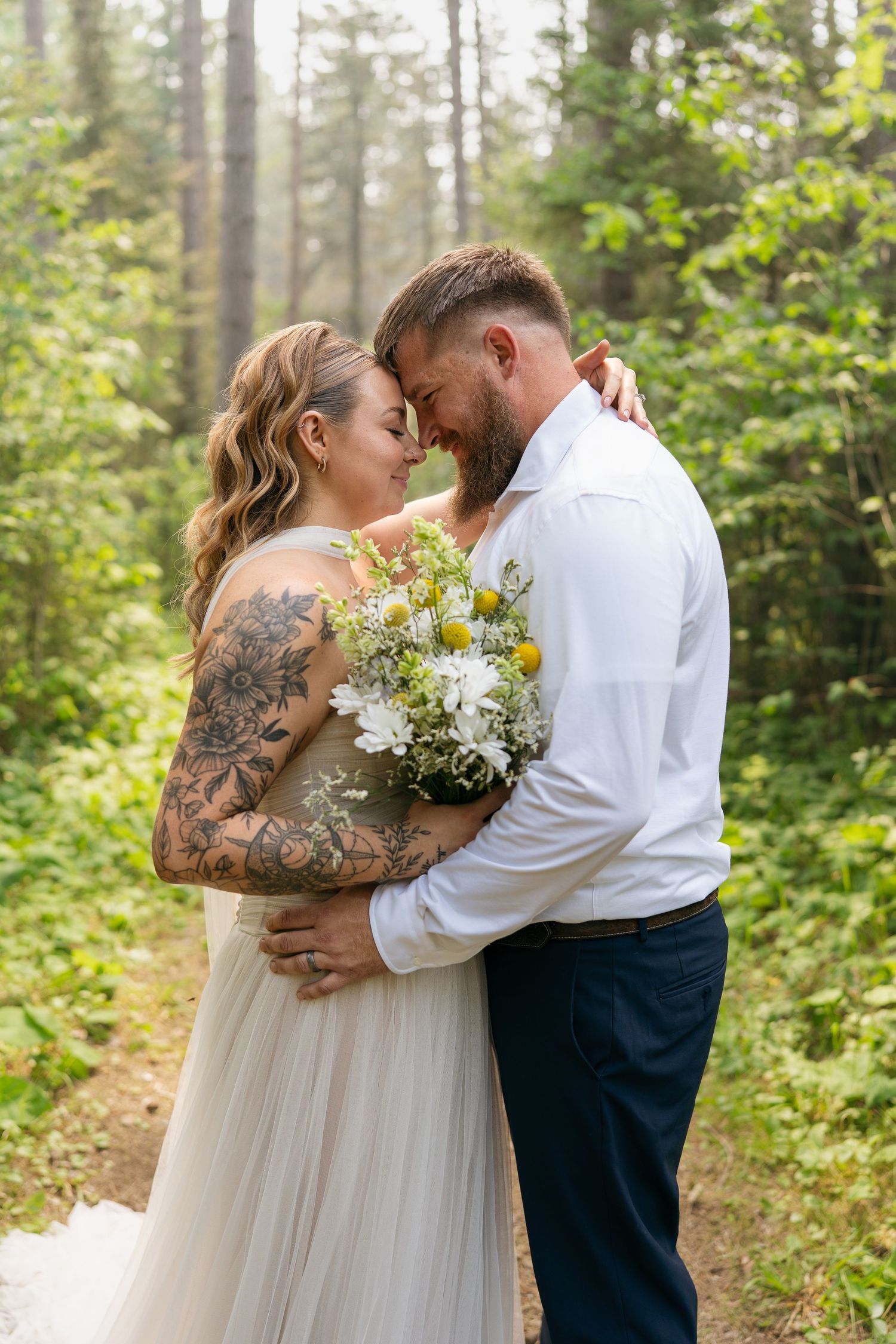 Tattooed bride and groom embrace in a misty forest, holding a wildflower bouquet in wedding attire.