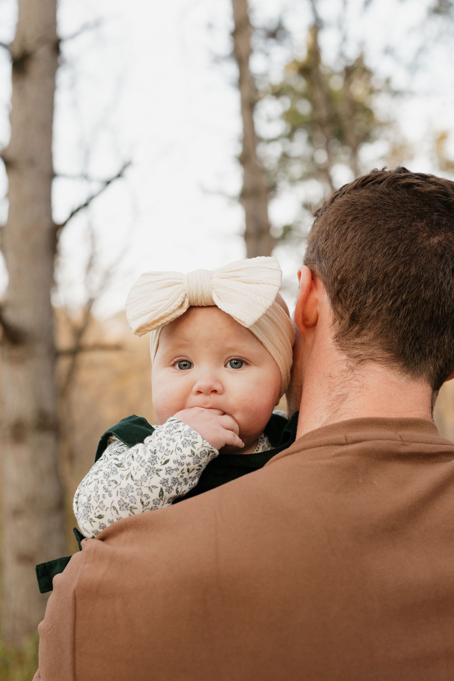 Close-up of curious baby girl with large cream bow headband resting on fathers shoulder outdoors.