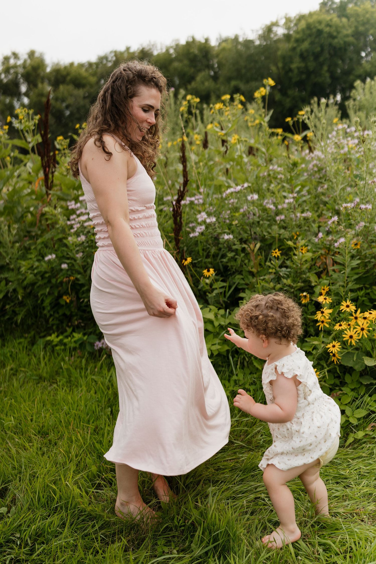 A mother in a pink dress plays with her curly-haired toddler in a lush garden filled with yellow wildflowers.