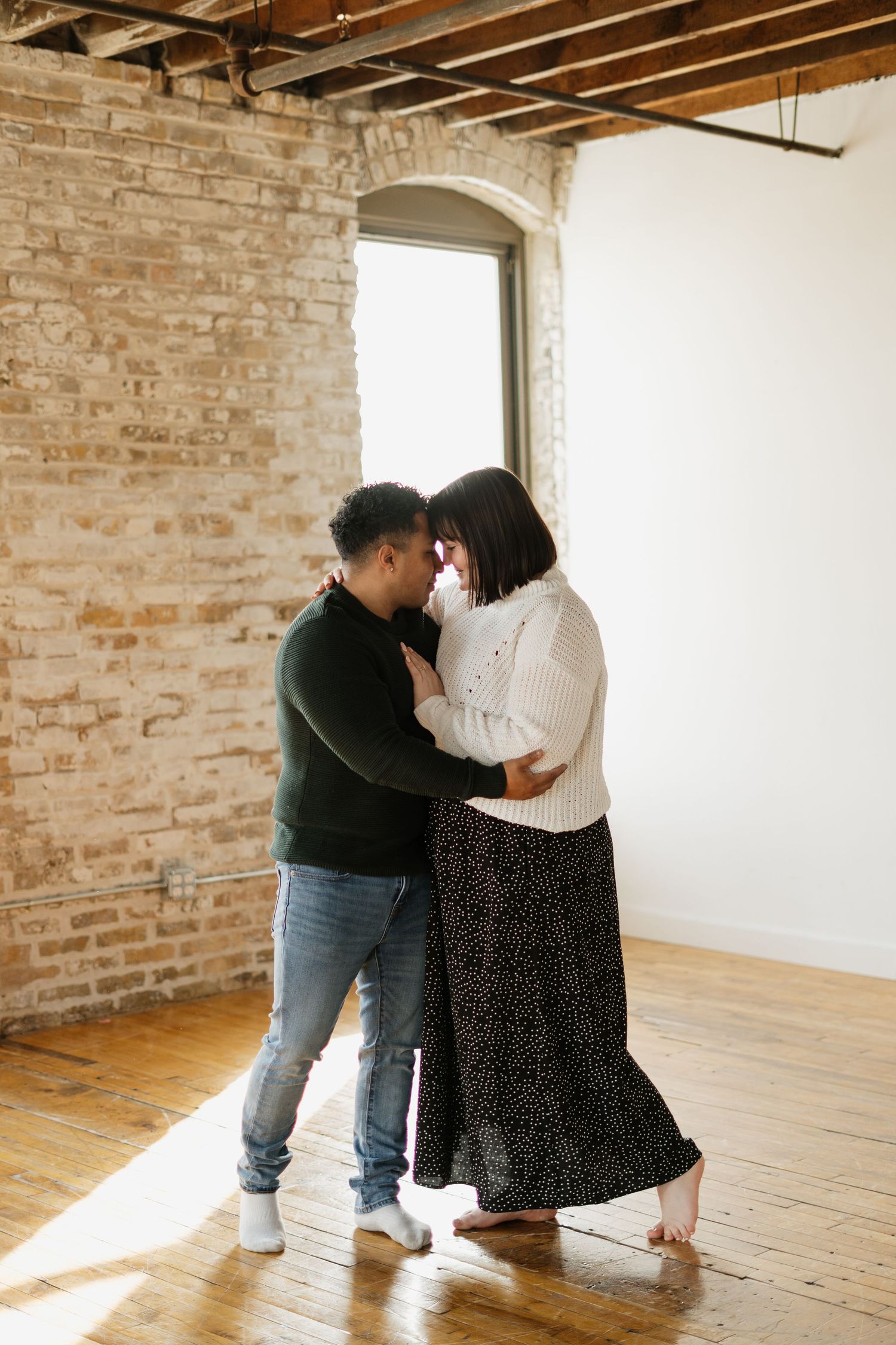 Couple sharing an intimate moment dancing in a rustic brick-walled studio with hardwood floors and natural window light.