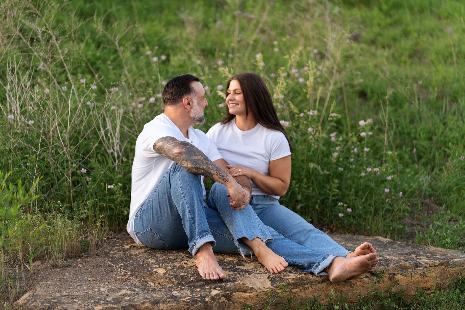 Couple sitting on a rock in a grassy field, wearing white tees and jeans, smiling at each other outdoors.