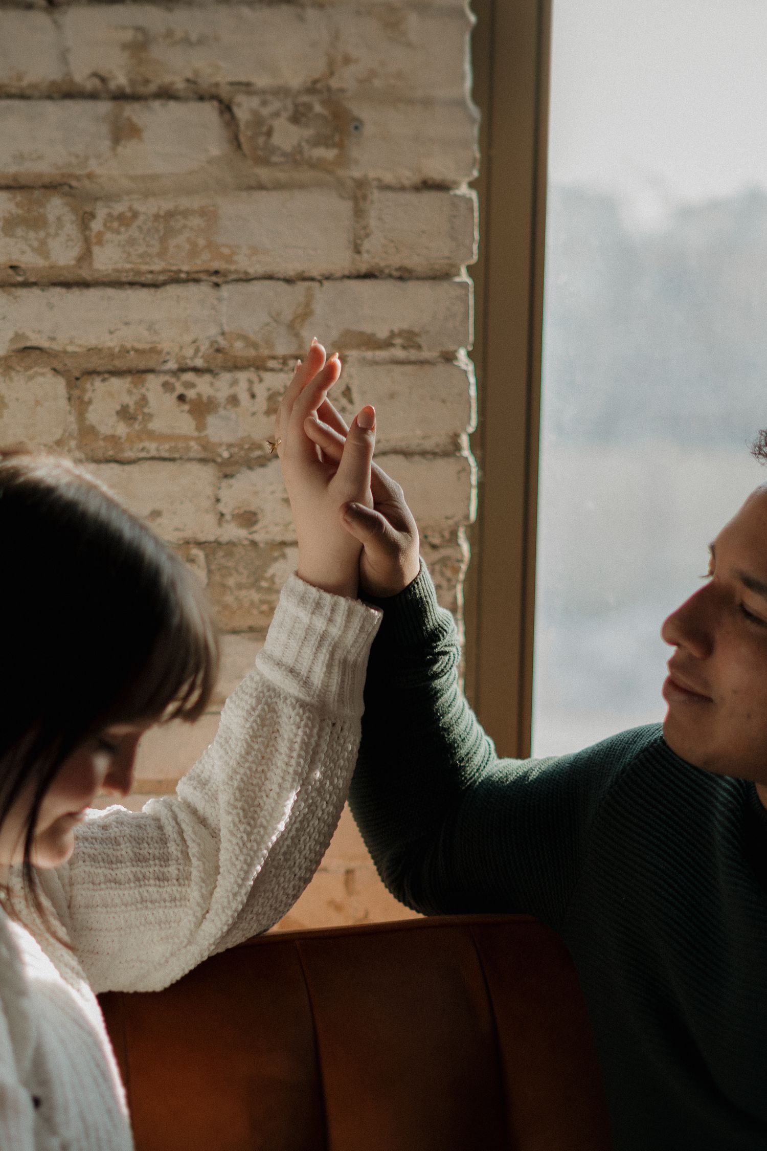 A man and woman in cozy sweaters hold hands near a brick wall window in a warm, intimate indoor setting.