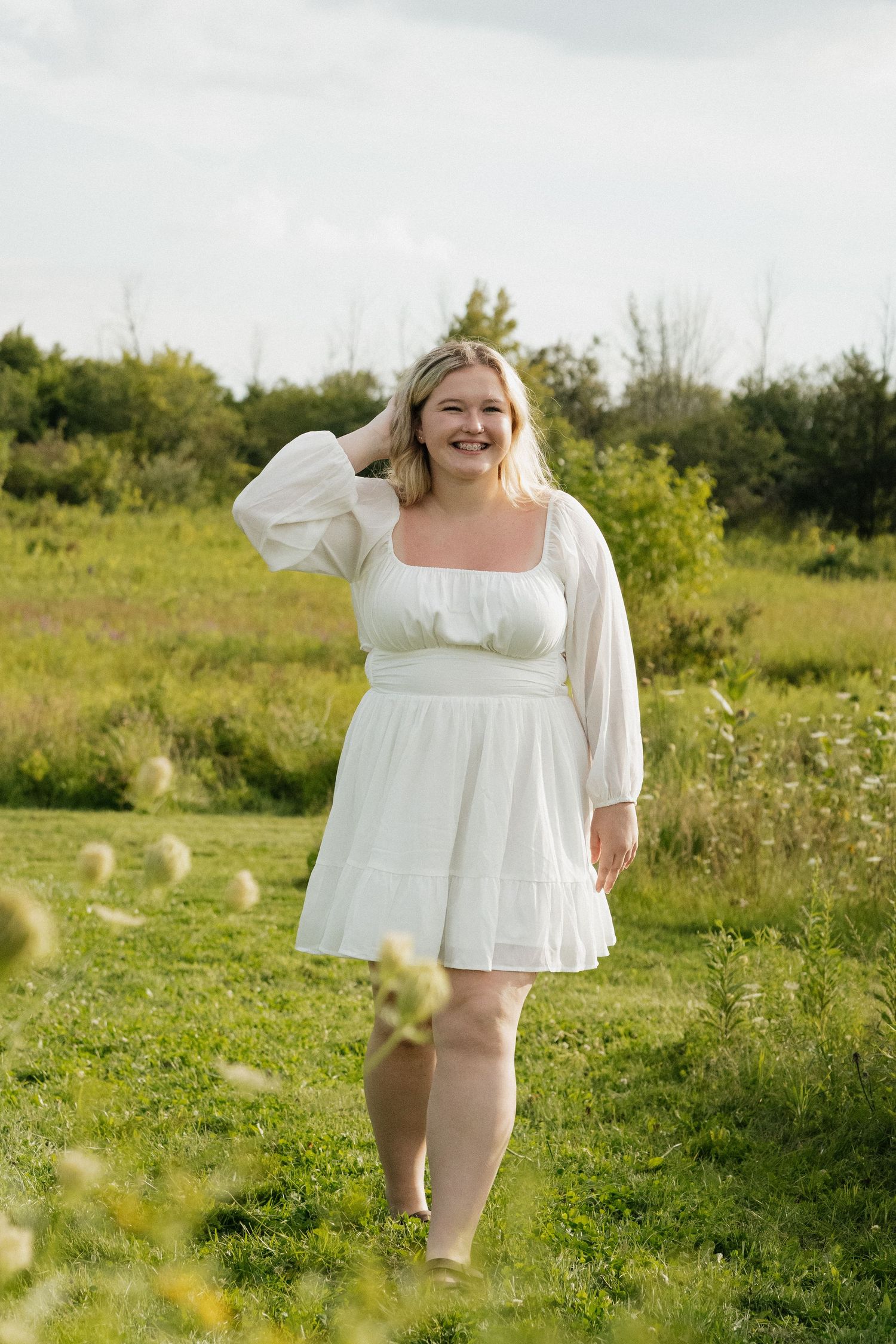 Smiling blonde woman in white dress posing in sunny green meadow.