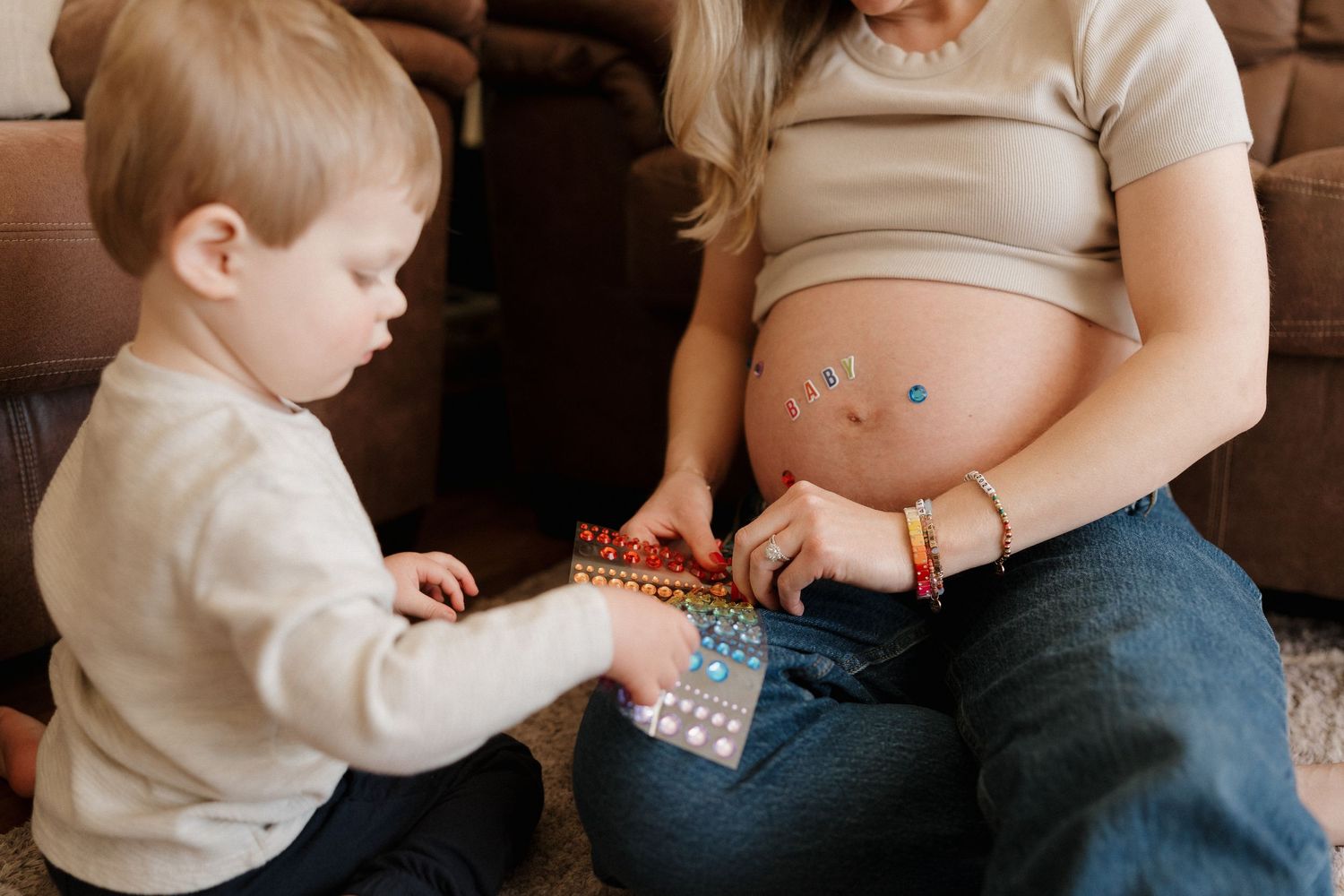 Pregnant woman with stickers on belly sits on floor with toddler child playing with colorful toy book.