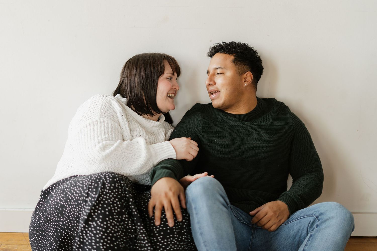 A happy couple sits on the floor laughing and talking together in a bright, cozy room.