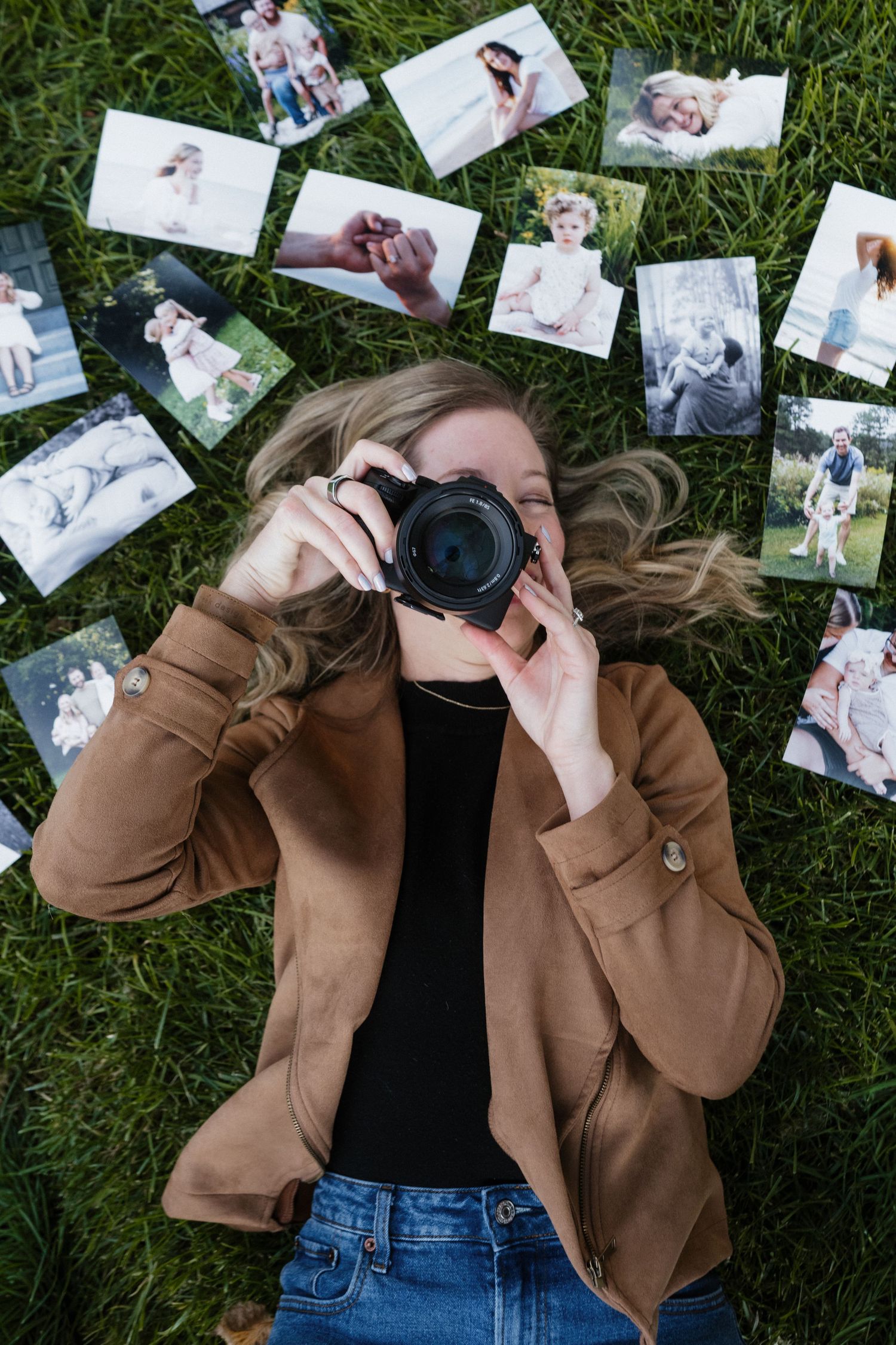 Woman lying on grass surrounded by printed photos, shooting upward with camera in brown jacket.