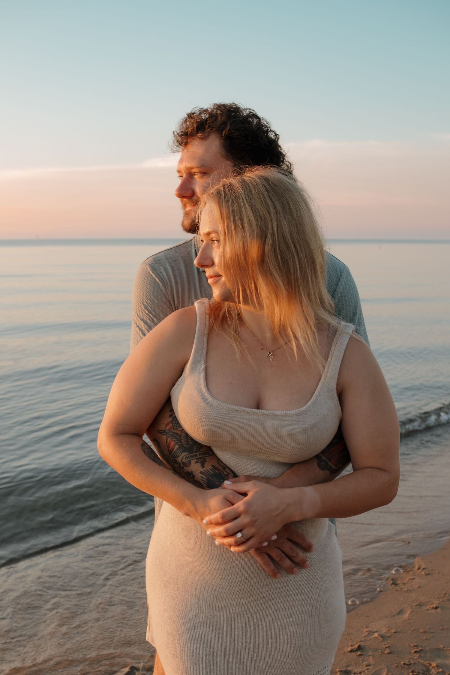 A couple shares a tender embrace on a beach at sunset, the woman in a knit dress leaning into the man.