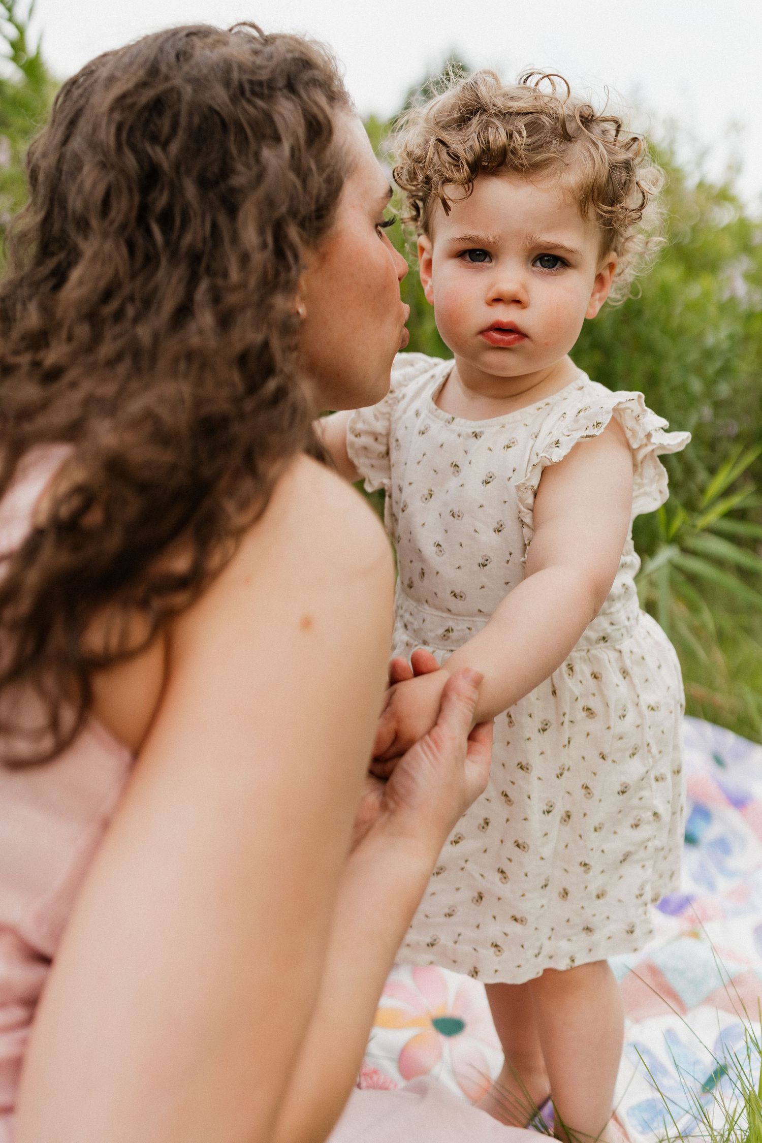 A curly-haired toddler in a white dress holds her mothers hand on a colorful outdoor blanket.