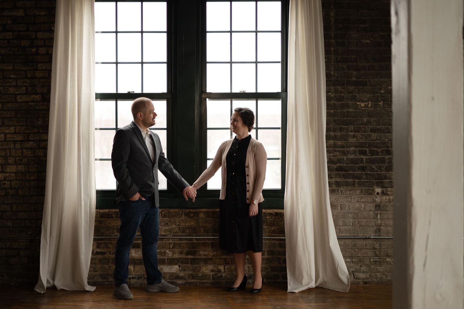 Couple holding hands near large windows with white curtains in a rustic brick-walled loft space.