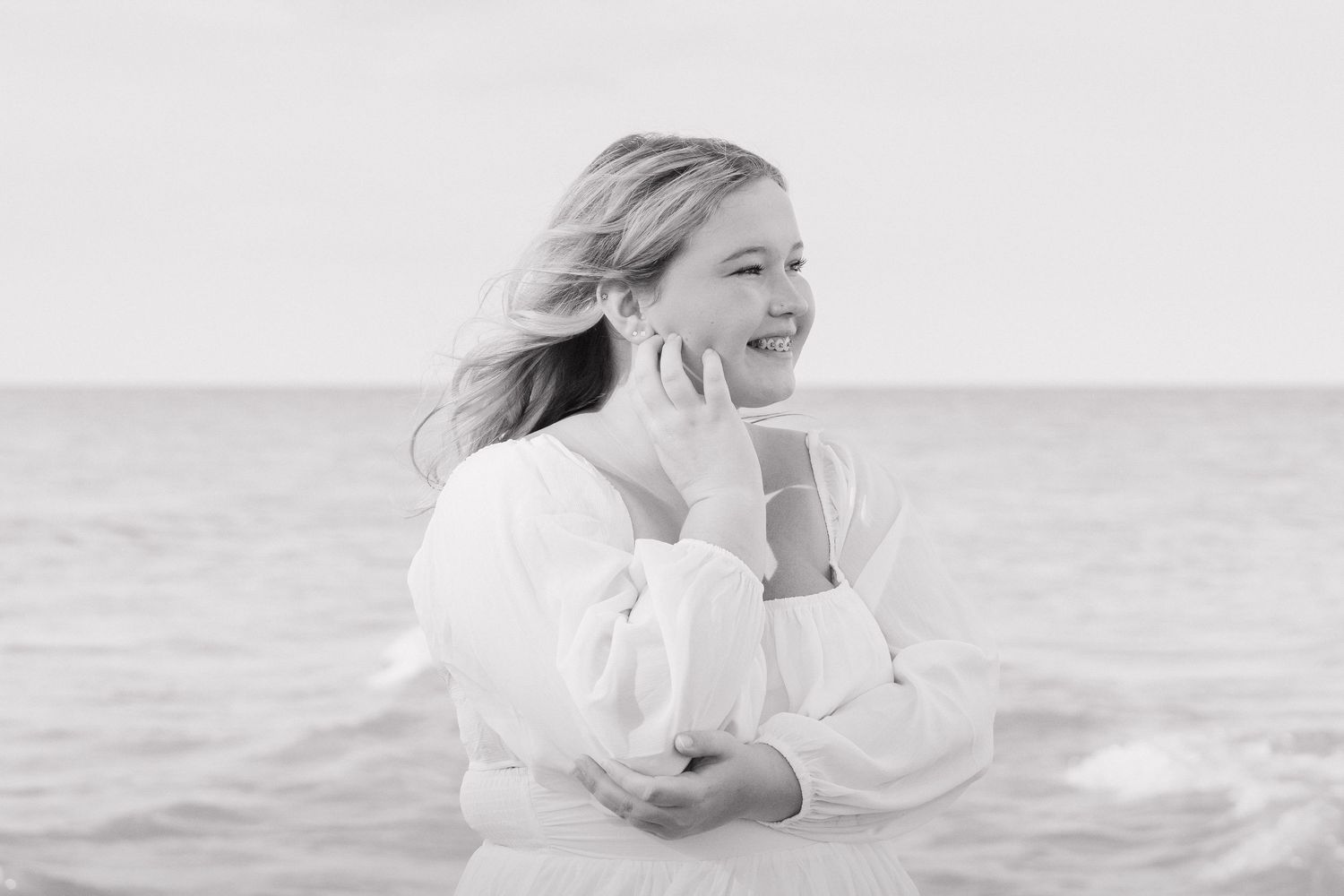Young blonde girl in white top smiles at ocean shore in black and white photo.