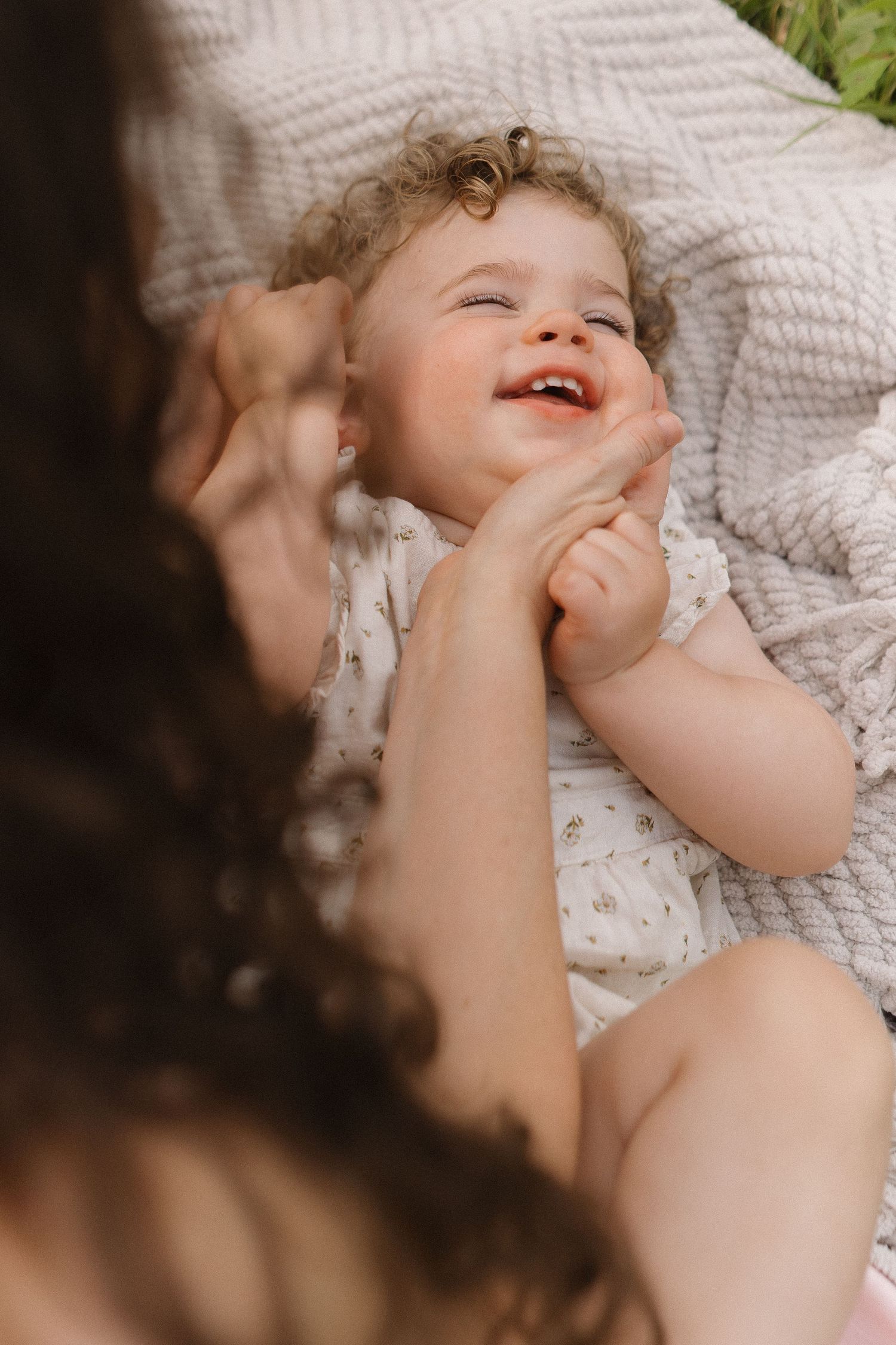 Joyful curly-haired baby girl laughs while lying on blanket as mother plays with her hands.