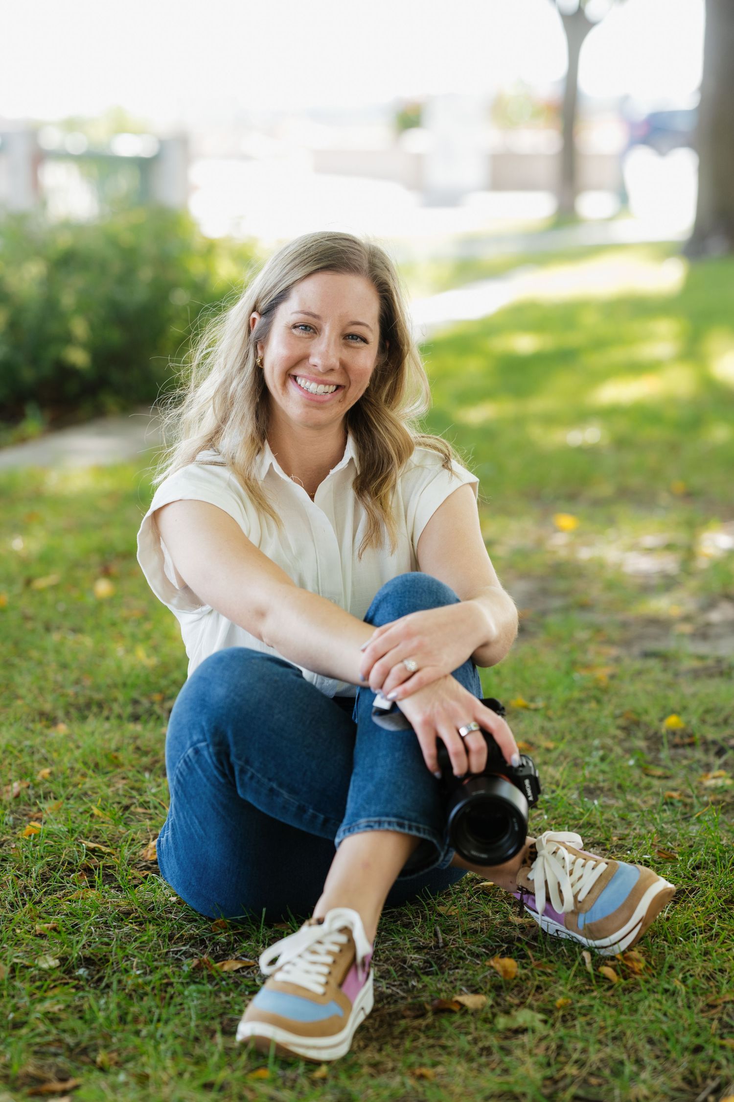 Smiling blonde female photographer sitting on grass outdoors, holding a camera, wearing jeans and white shirt.