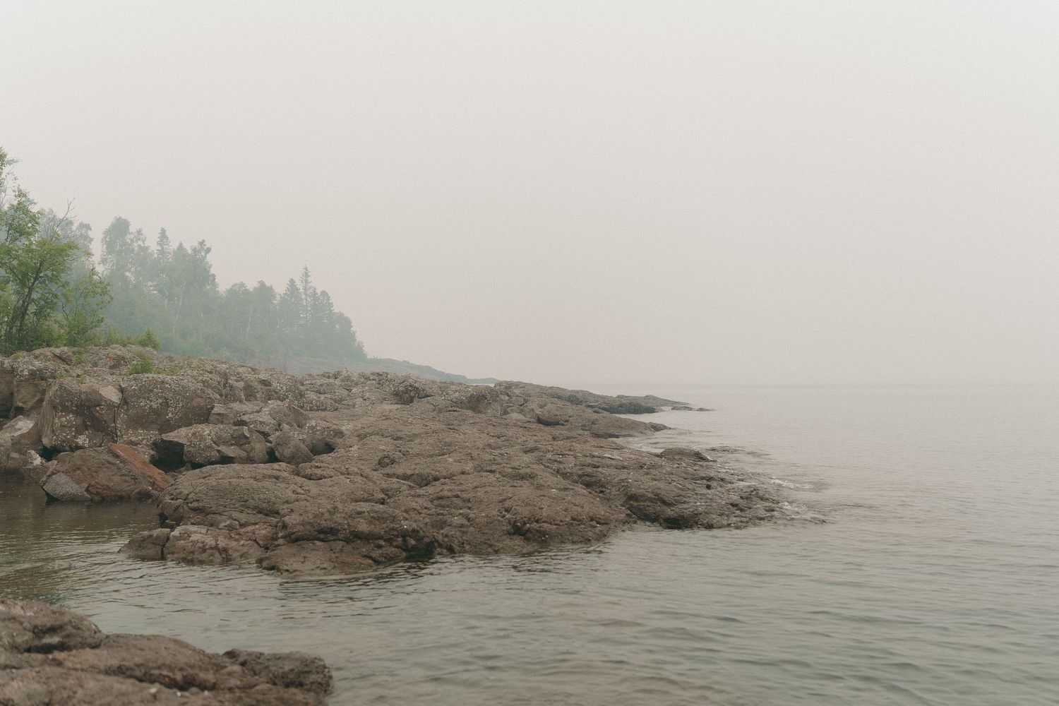 Misty rocky shoreline with calm water and fog-covered trees in the background on a grey overcast day.