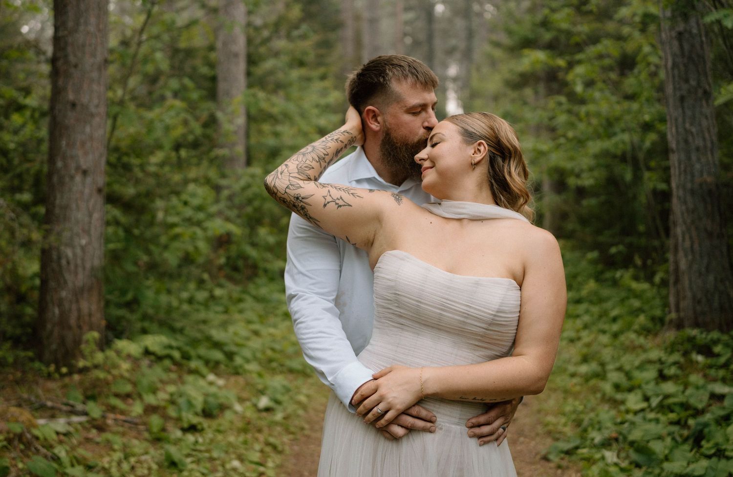 Couple sharing a romantic moment in a lush forest, groom with tattoos embracing bride in a strapless gown.