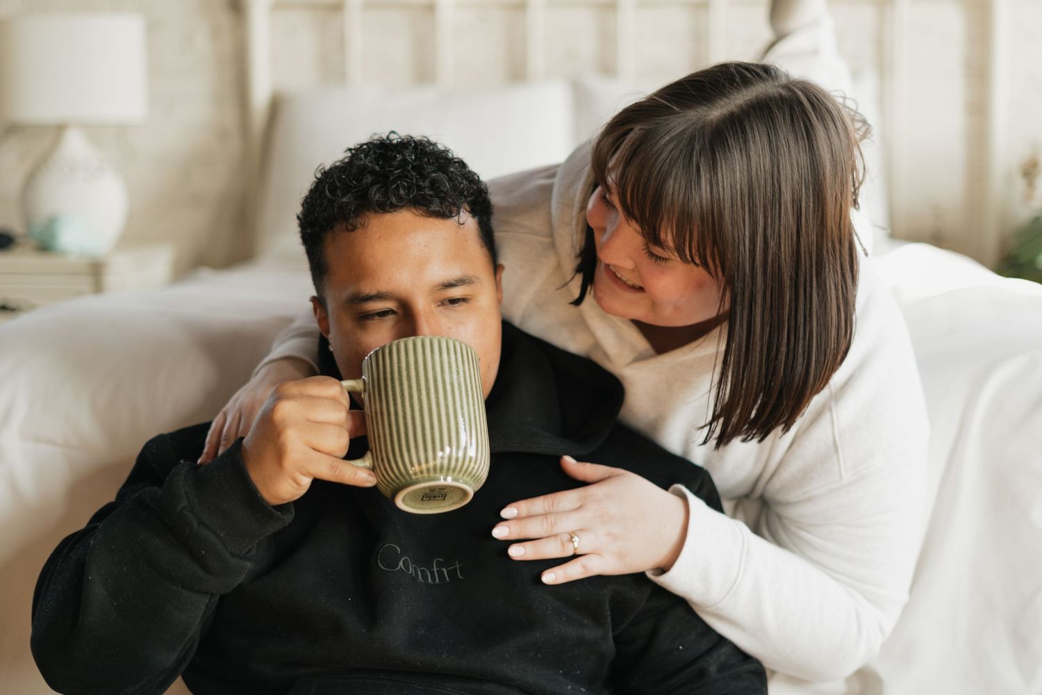 Couple relaxing in bedroom, man drinking from green mug while woman embraces him from behind.