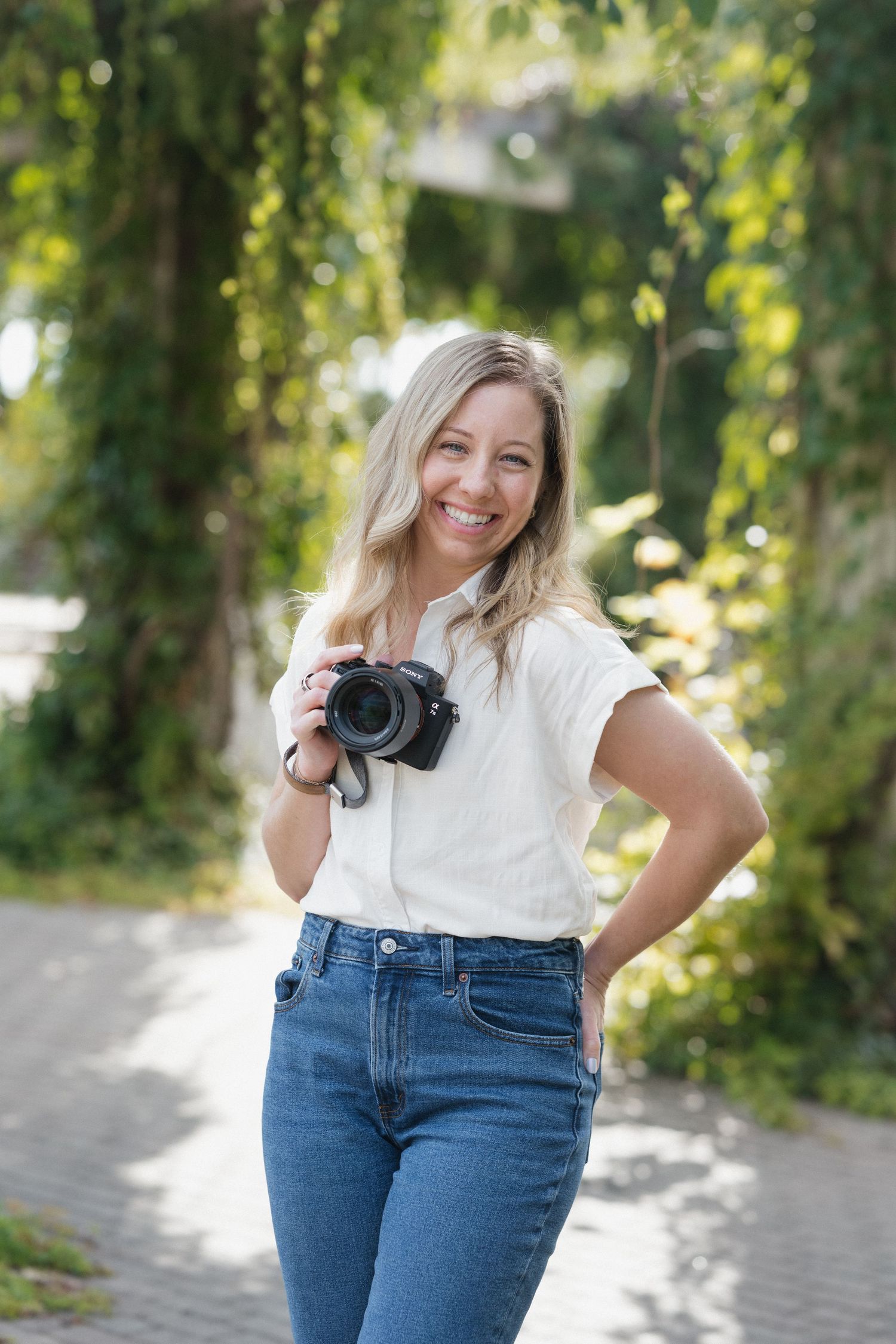 Smiling blonde female photographer holds camera outdoors surrounded by lush green foliage.