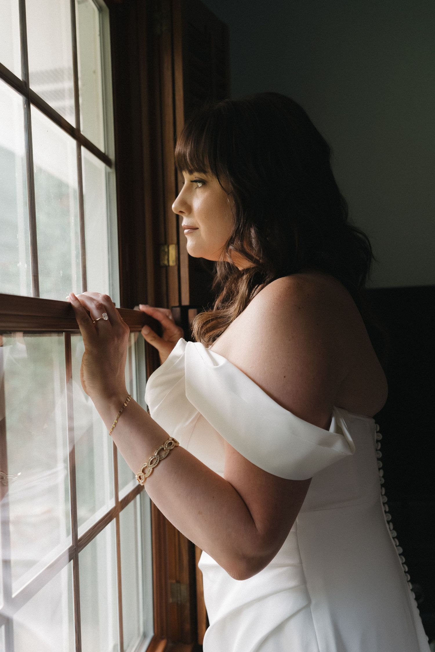 Bride in off-shoulder white gown gazing thoughtfully out a window on her wedding day.