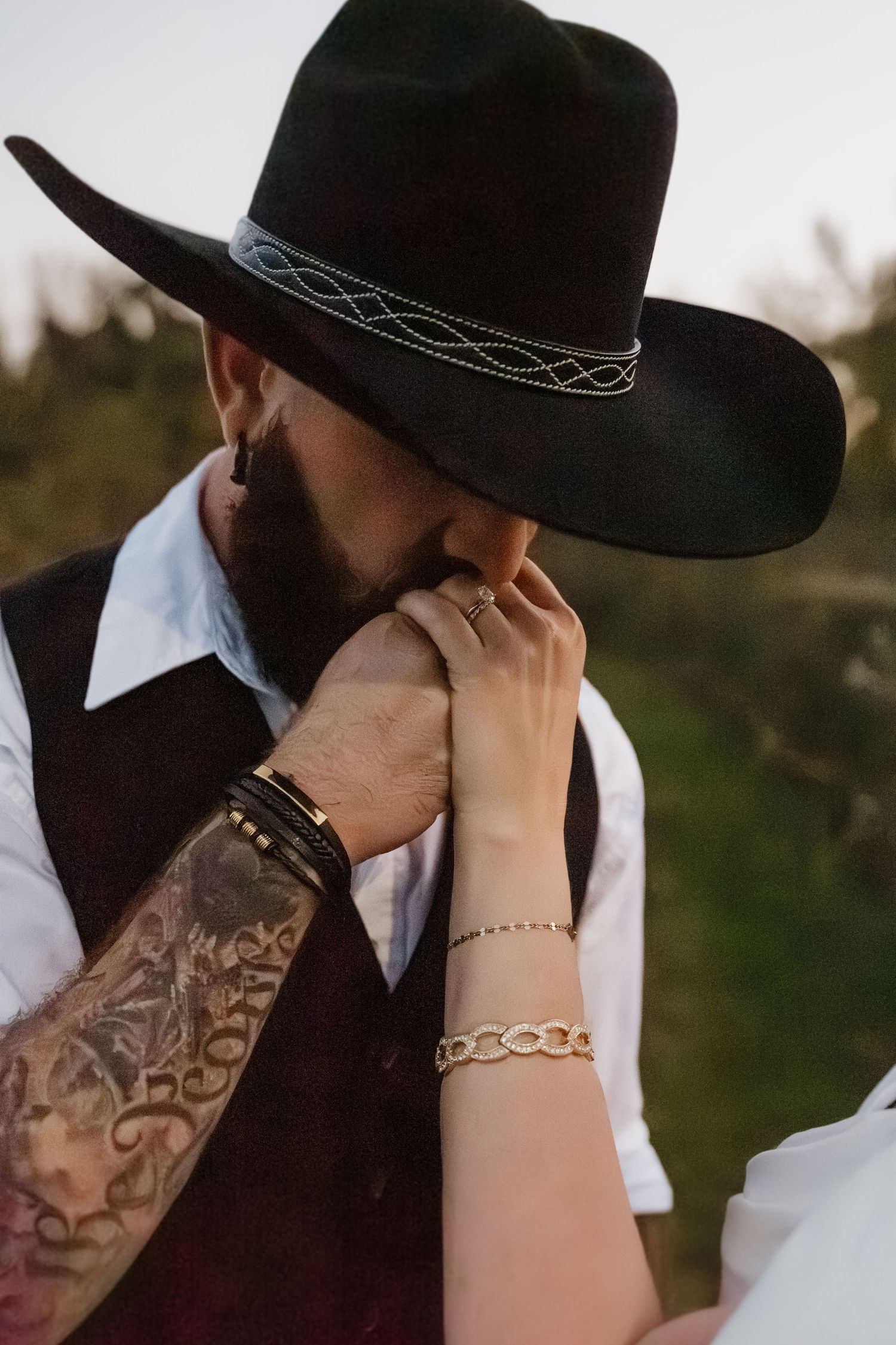 Tattooed groom in black cowboy hat kisses bride's hand adorned with gold bracelet outdoors.