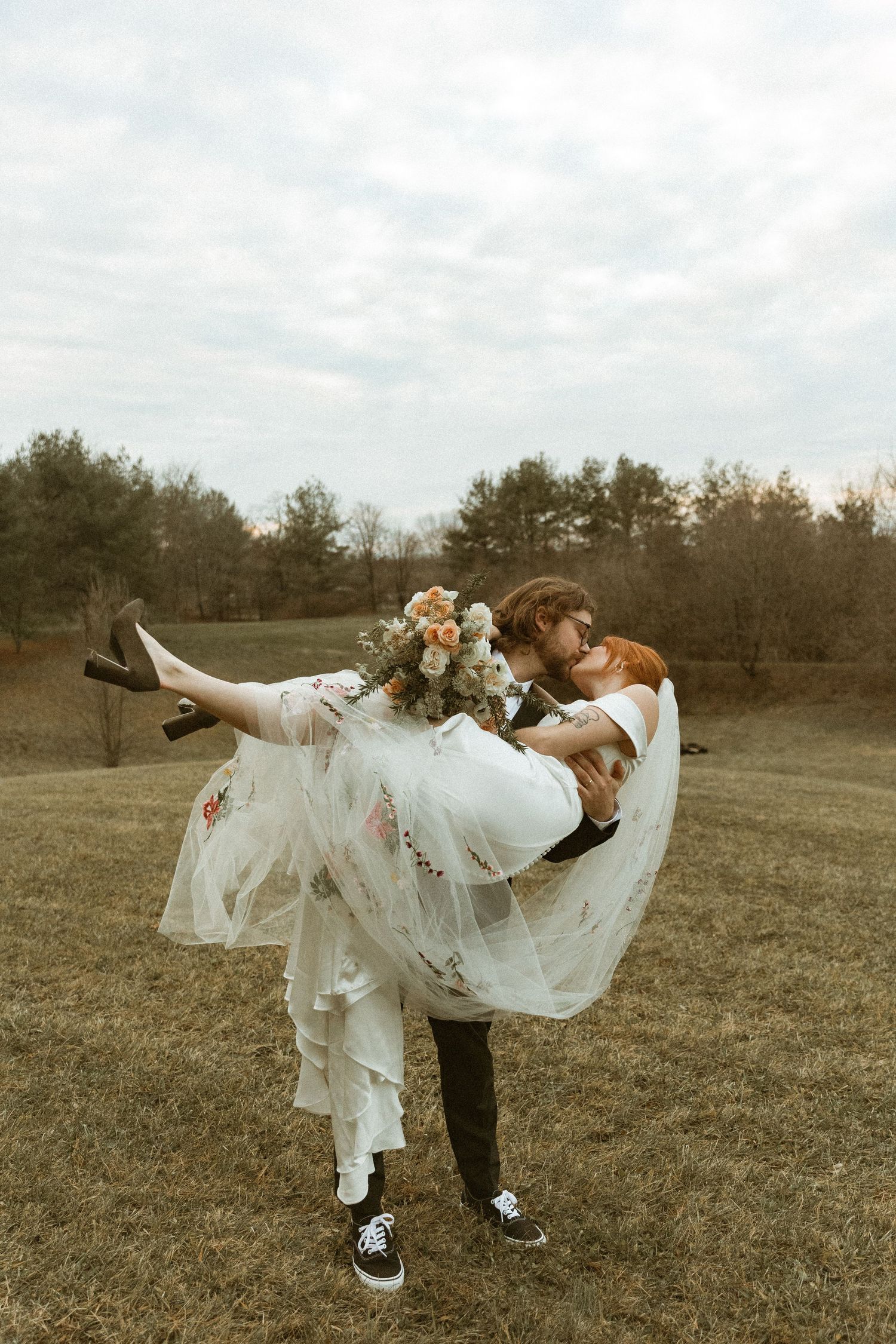 Playful wedding portrait of groom lifting bride in white dress and sneakers in an open field.