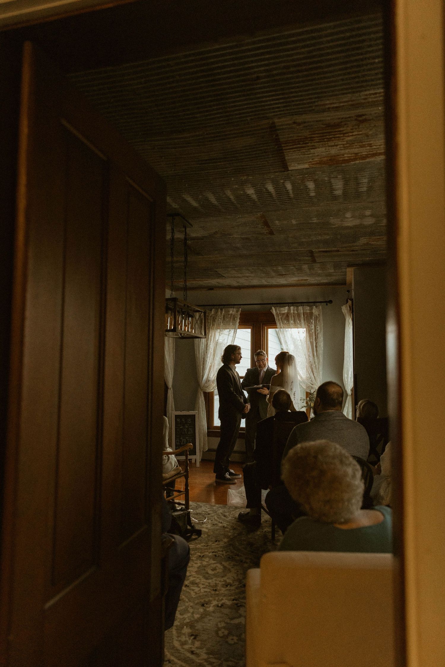 An intimate wedding ceremony viewed through a doorway with guests seated in a rustic indoor setting.