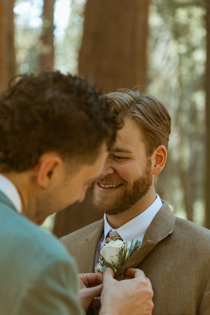 A tender moment captured as one person adjusts the other's boutonniere in a forest setting.