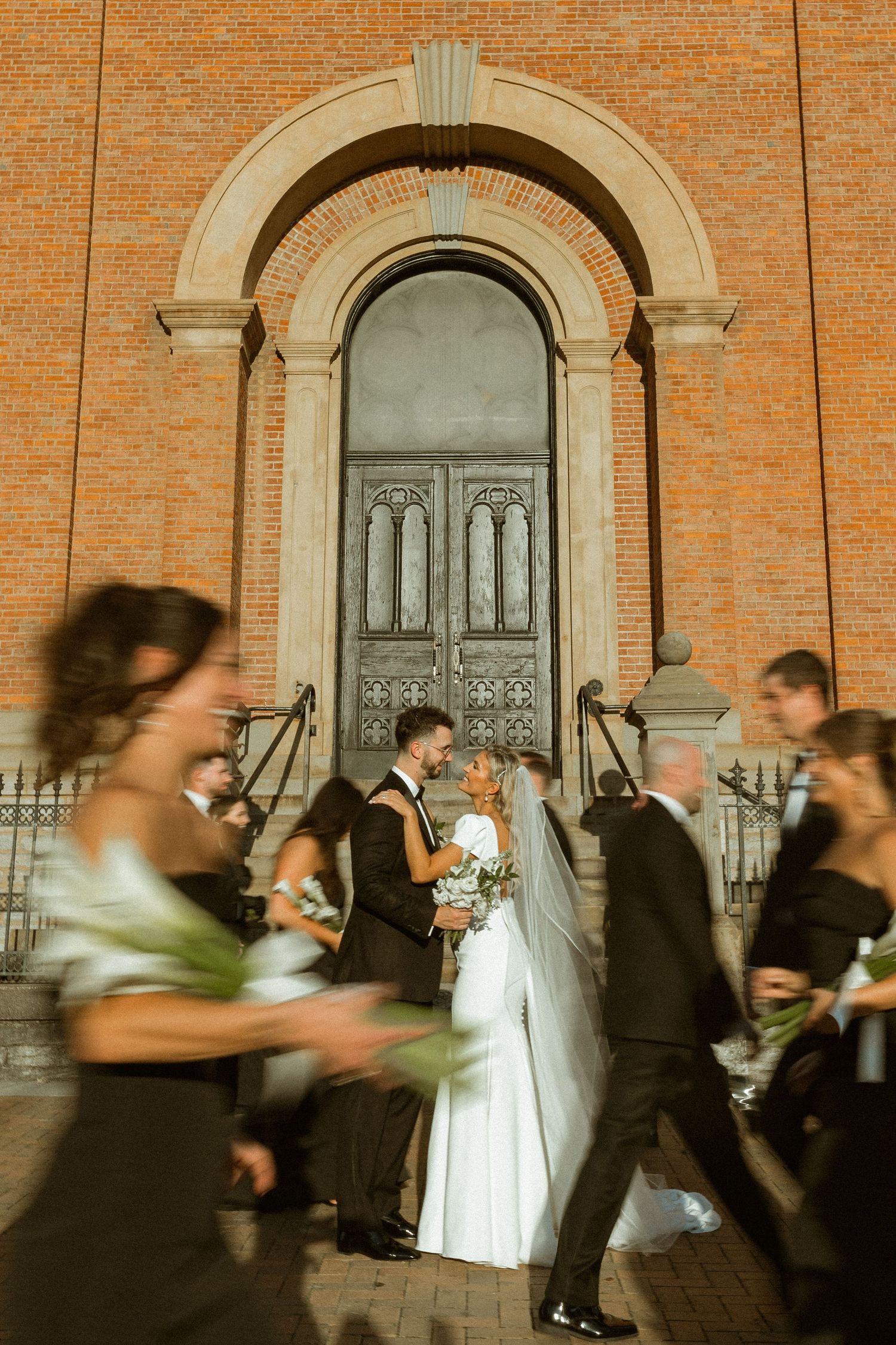 Motion blur of wedding party and guests outside brick building with arched doorways.