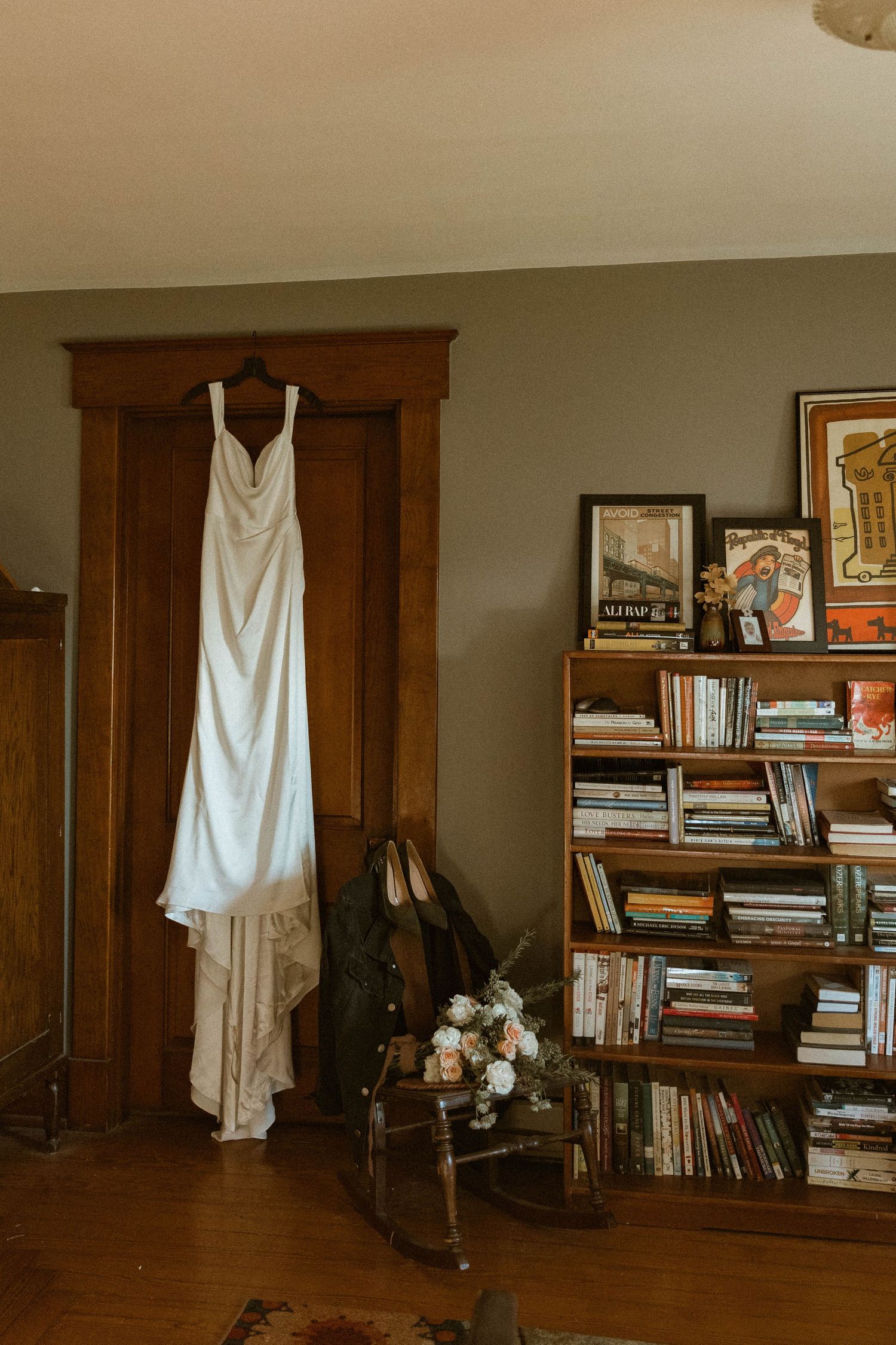A white wedding dress hangs elegantly in front of a wooden bookshelf with vintage decor.