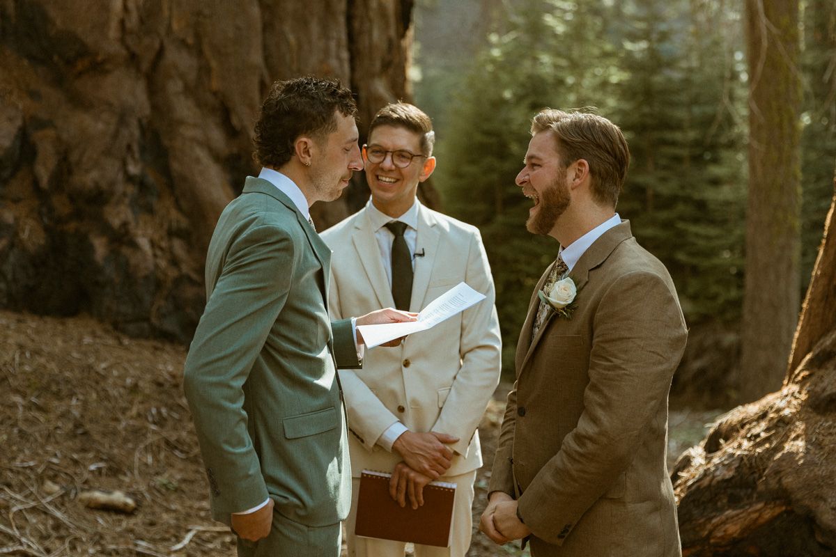 An outdoor wedding ceremony in a forest with people in formal attire gathered among tall trees.