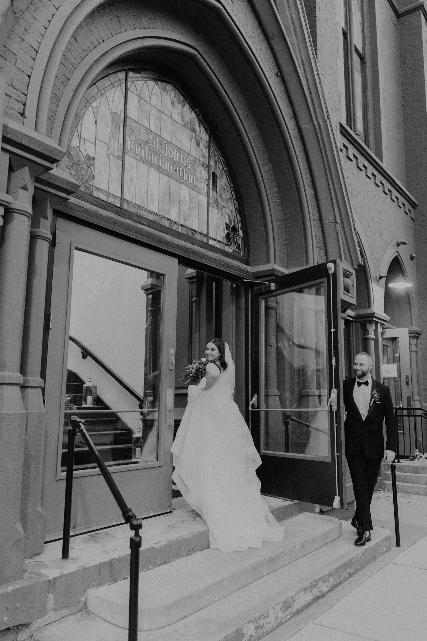 A black and white wedding photo of a couple exiting an arched doorway of a historic building.