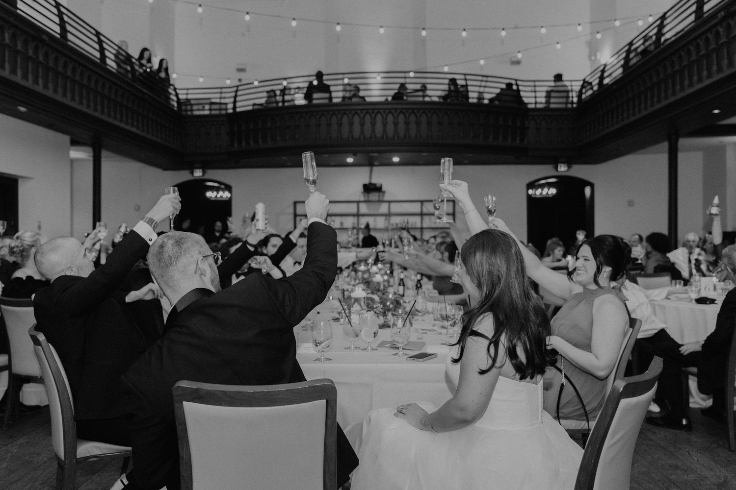 Black and white photo of wedding reception dinner with guests raising their glasses in celebration.