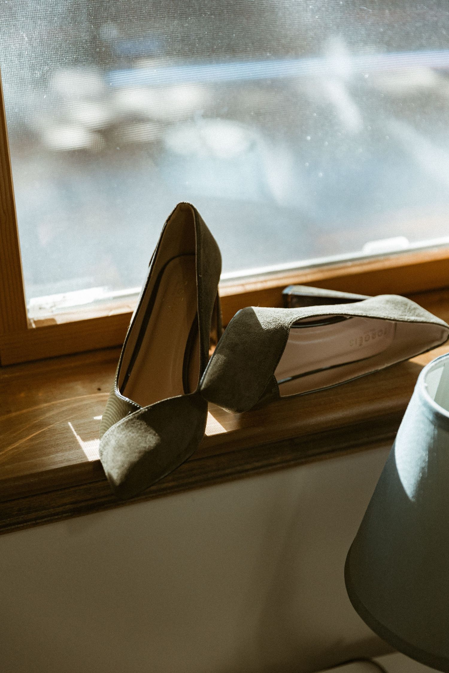 A pair of elegant grey wedding heels sits on a wooden windowsill in natural light.