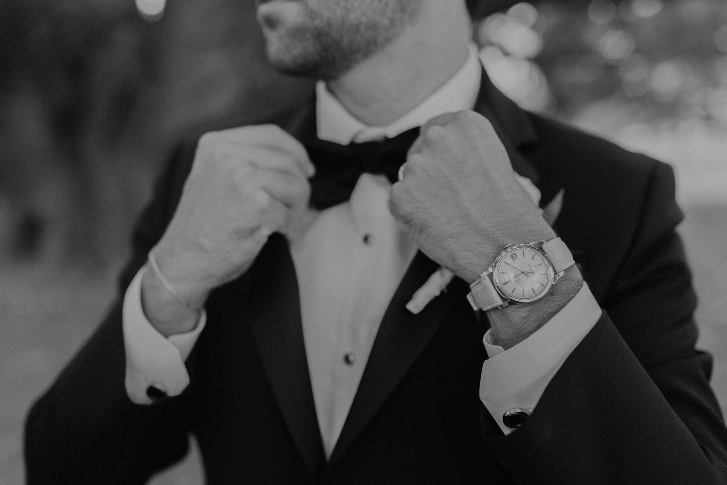 Close-up of groom adjusting bowtie while wearing luxury watch and tuxedo.