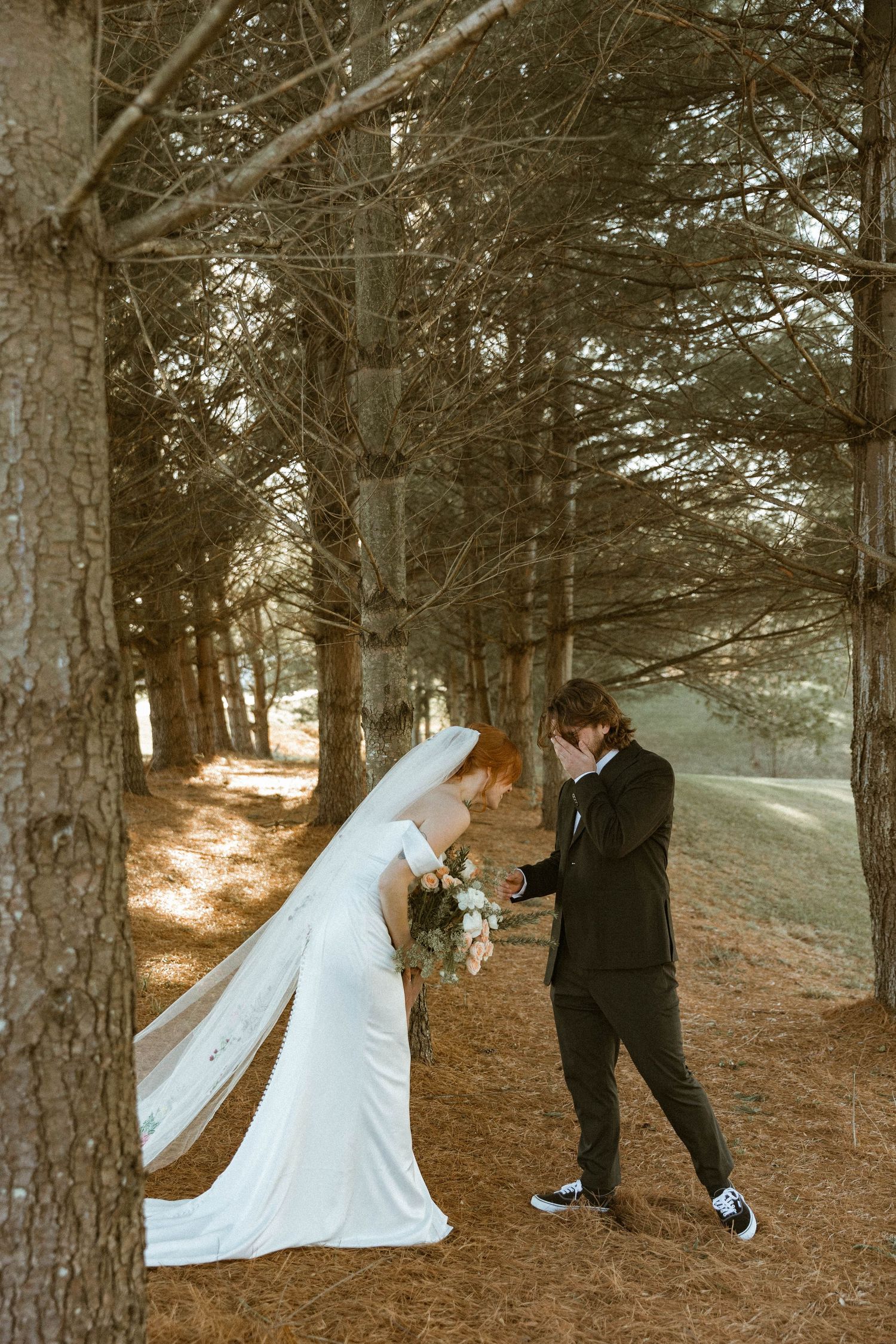 A couple in wedding attire shares a playful moment among tall pine trees.