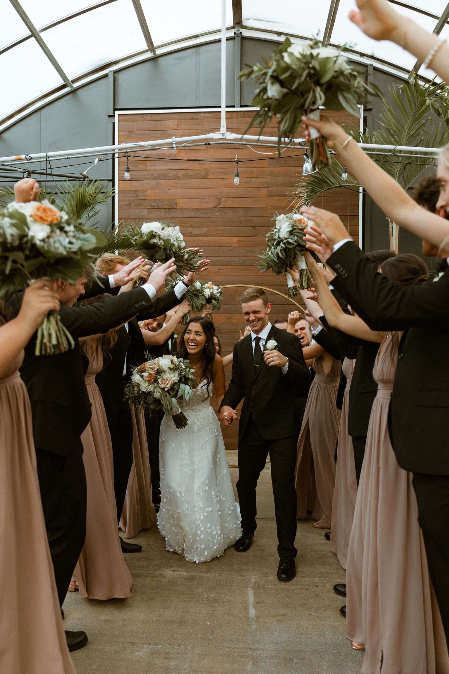 Wedding party in brown dresses and black suits poses with raised arms in celebration.