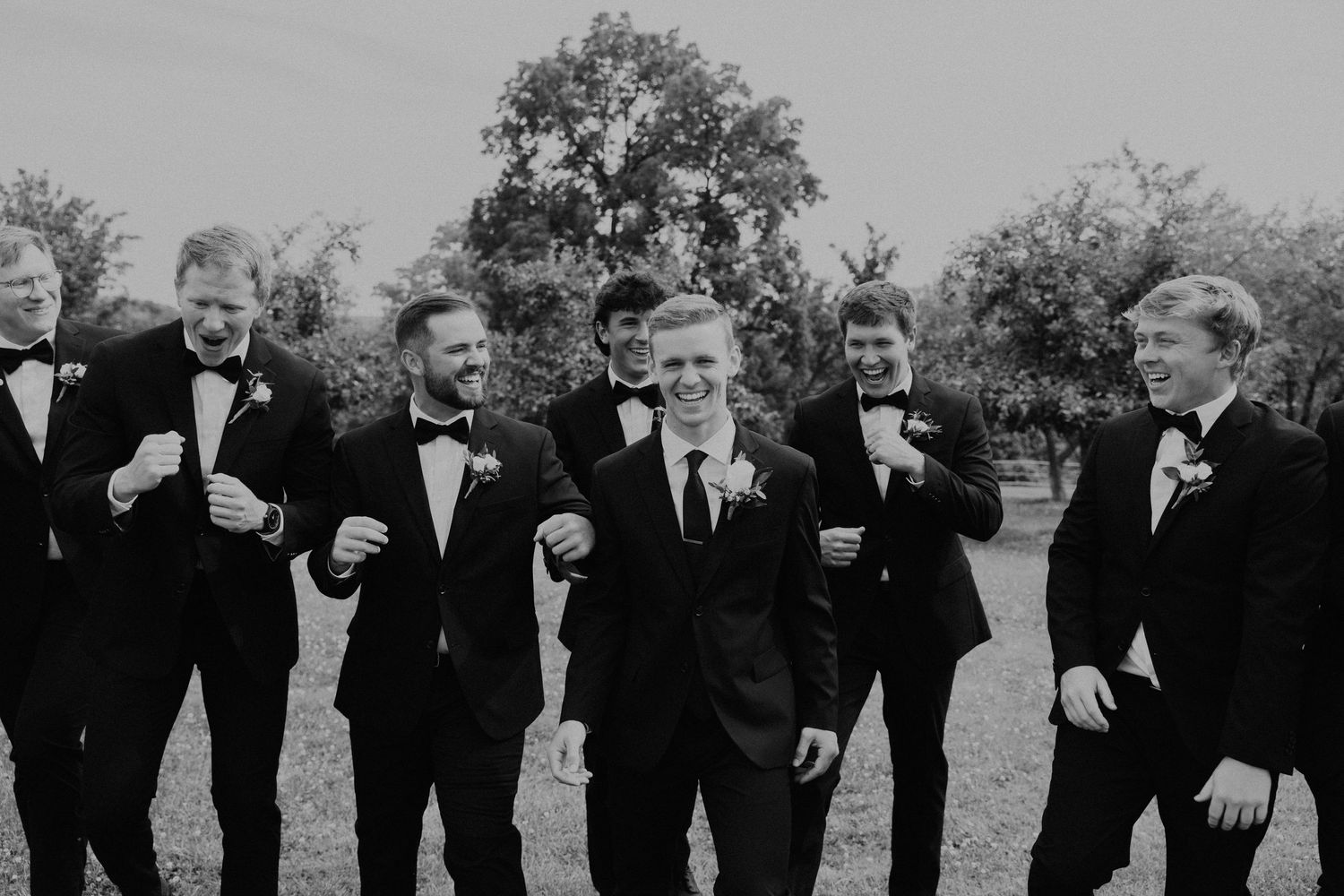 Groomsmen in black tuxedos line up for a candid black and white photo.