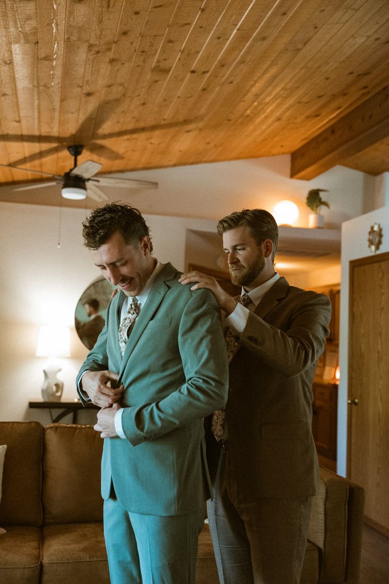 Two people in formal attire helping each other get ready in a room with wooden ceiling and warm lighting.