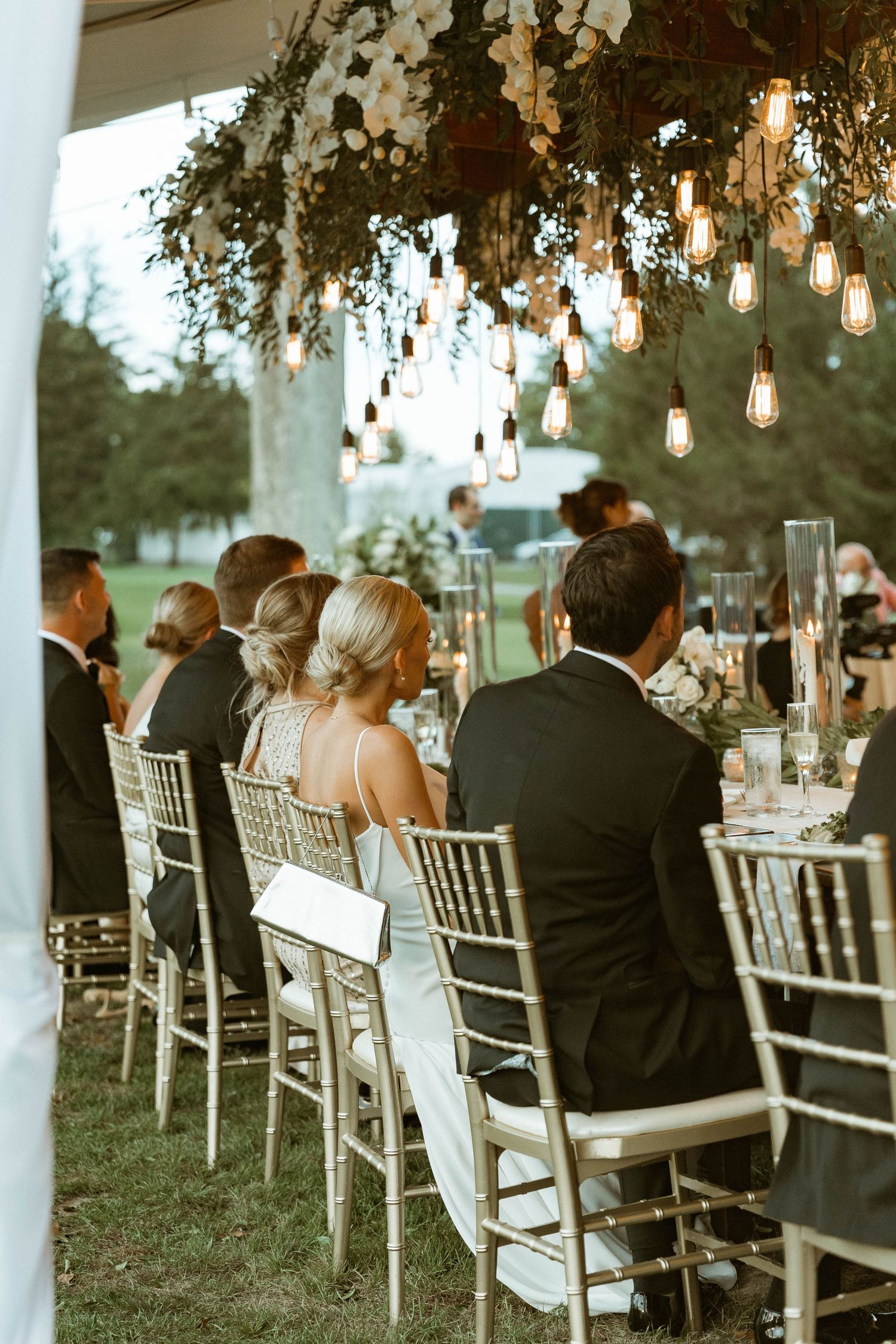 Elegant outdoor reception with hanging lights and floral arrangements above white chairs.