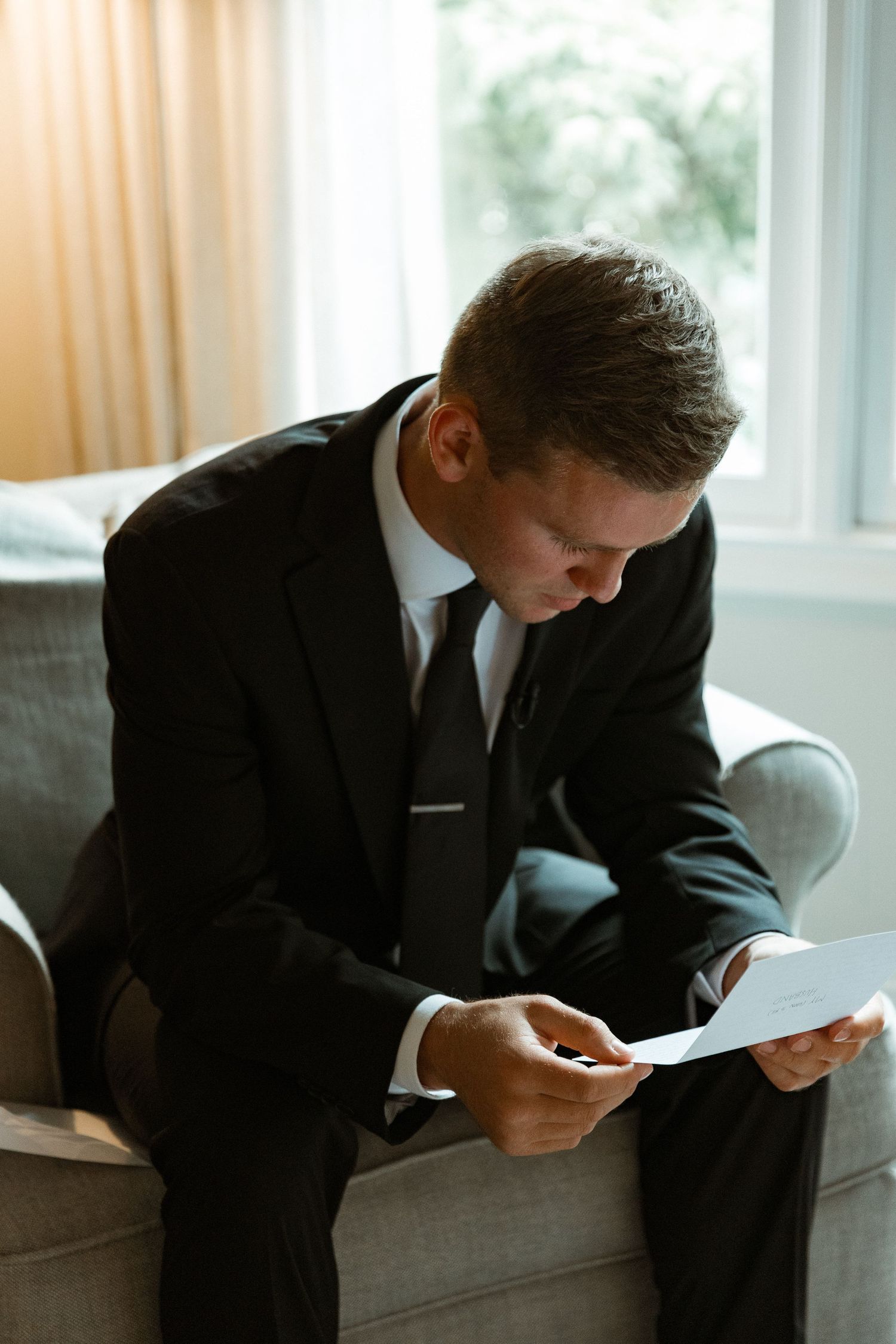Groom in black suit reading a letter while sitting on couch by window.