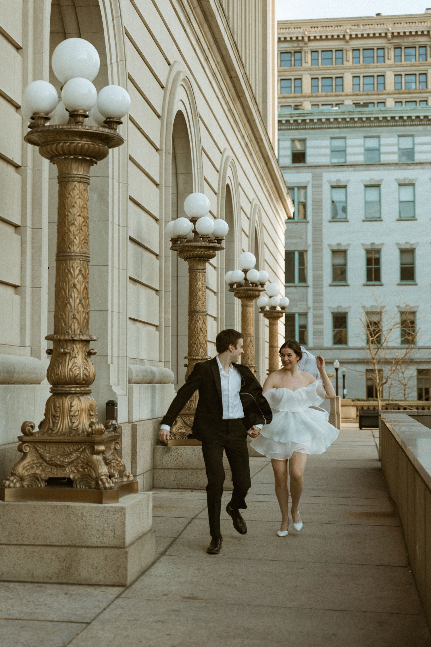 Couple running playfully along a classical building facade with ornate street lamps.