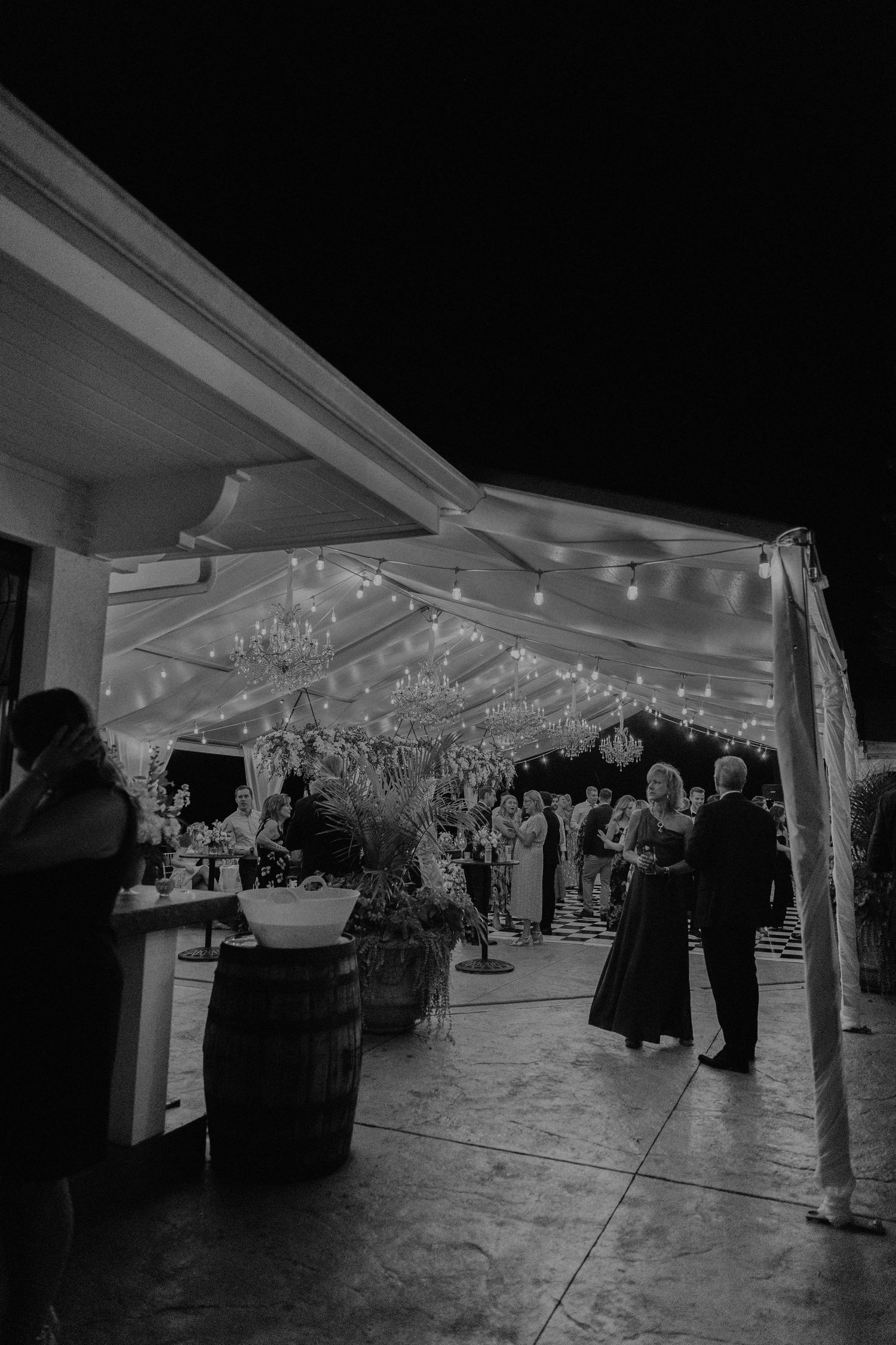 Black and white photo of wedding reception under tent with string lights and chandeliers.